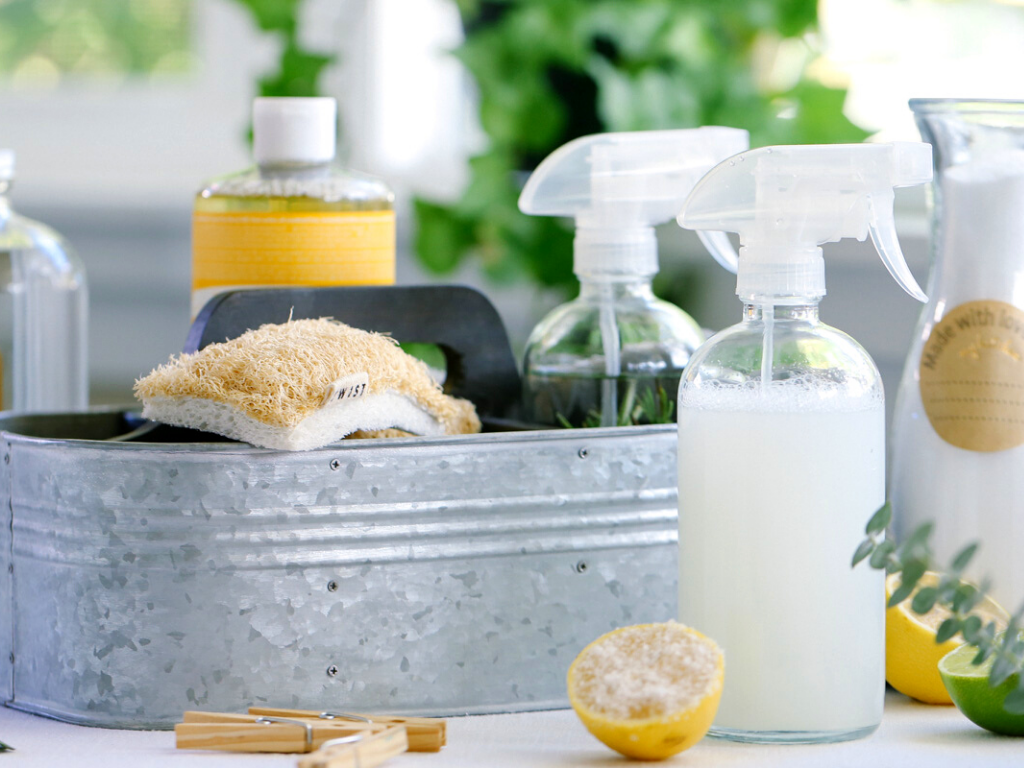 Cleaning supplies on a metal container, including spray bottles with cleaning solutions, a natural sponge, a lemon half, and other cleaning products, with greenery in the background.