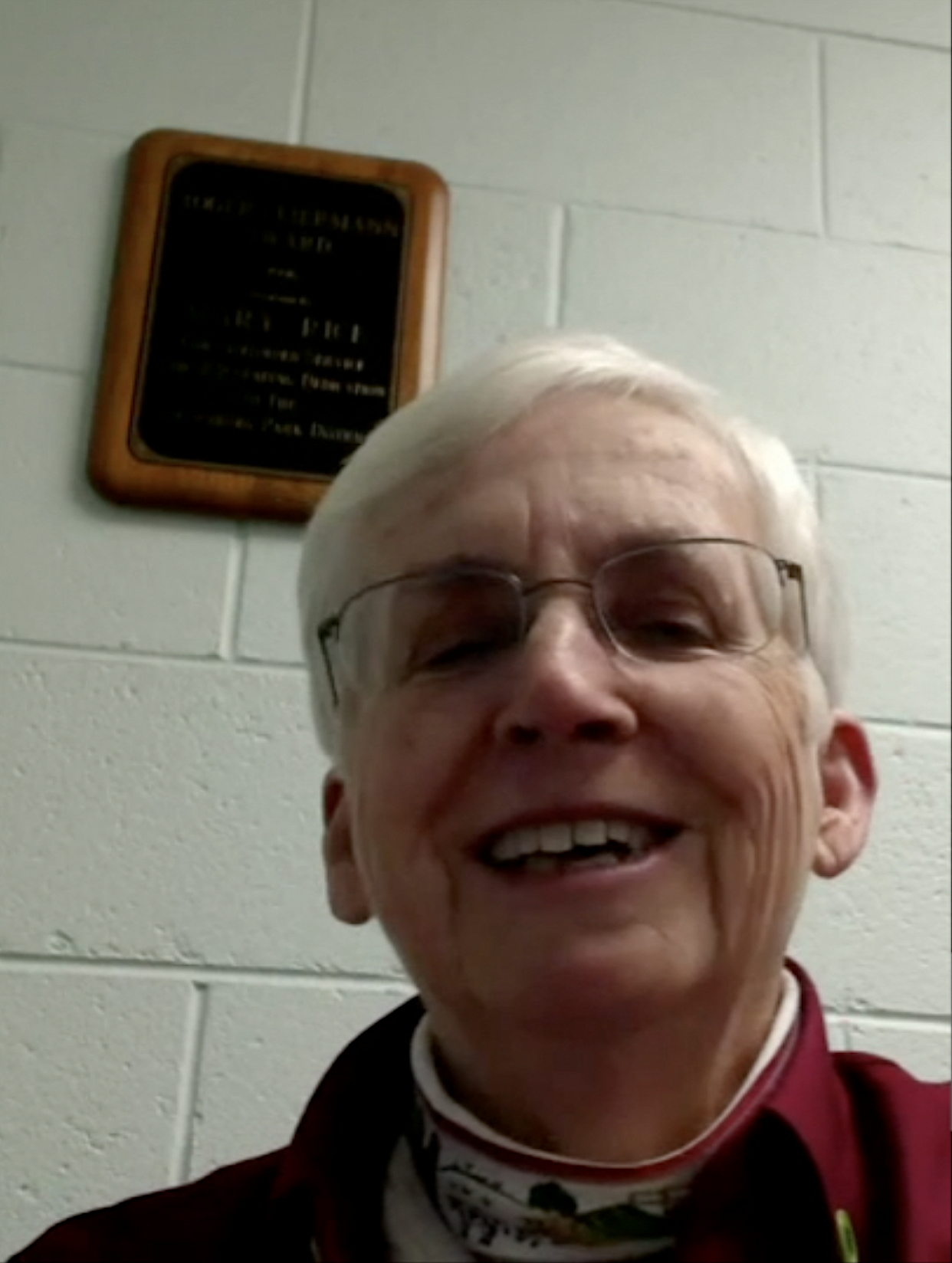 A smiling elderly man wearing glasses and a maroon shirt, standing in front of a white brick wall with a black and gold plaque mounted on it.