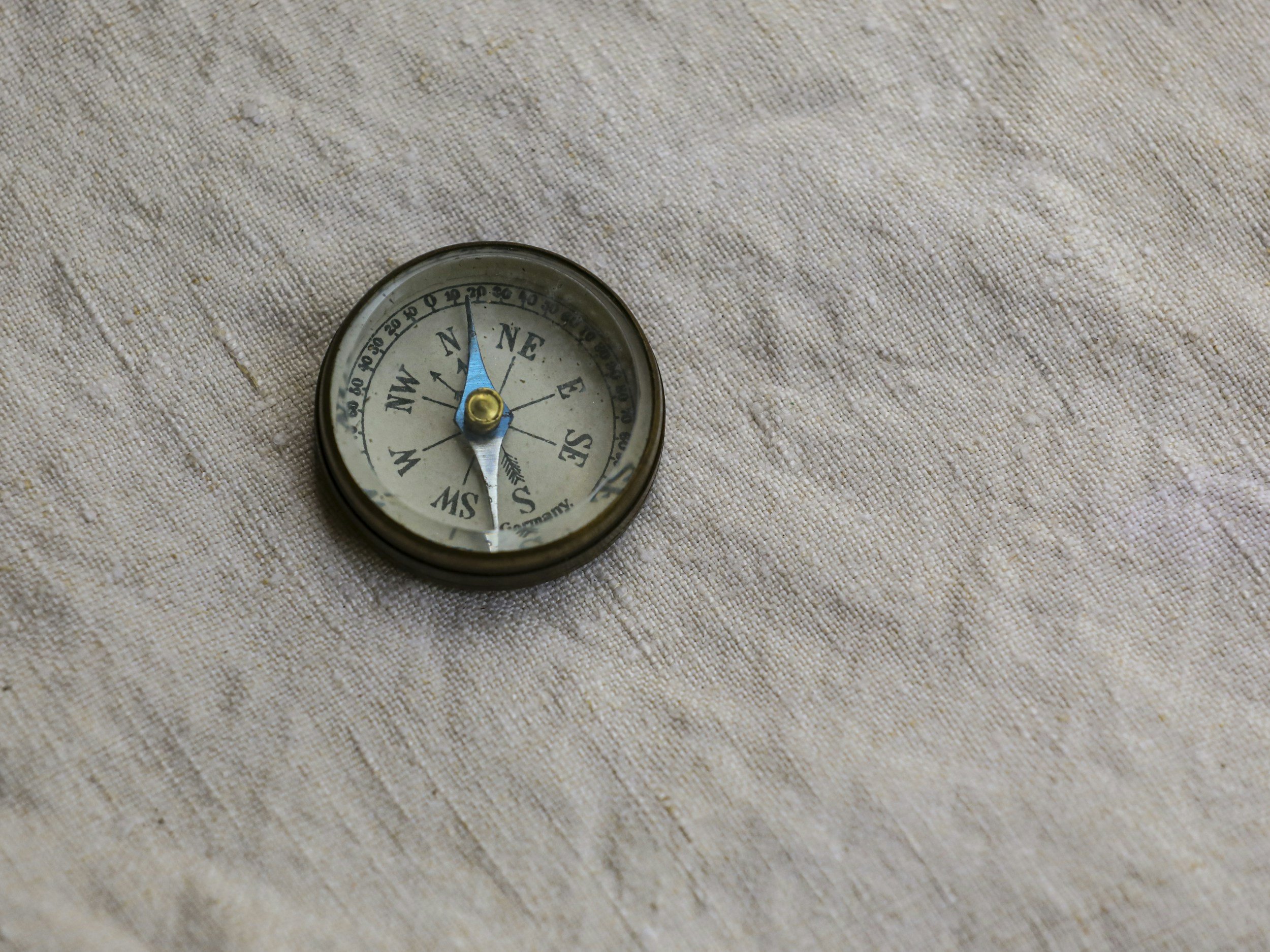 A small round compass with a brass pin on a textured beige fabric surface, showing north at the top.