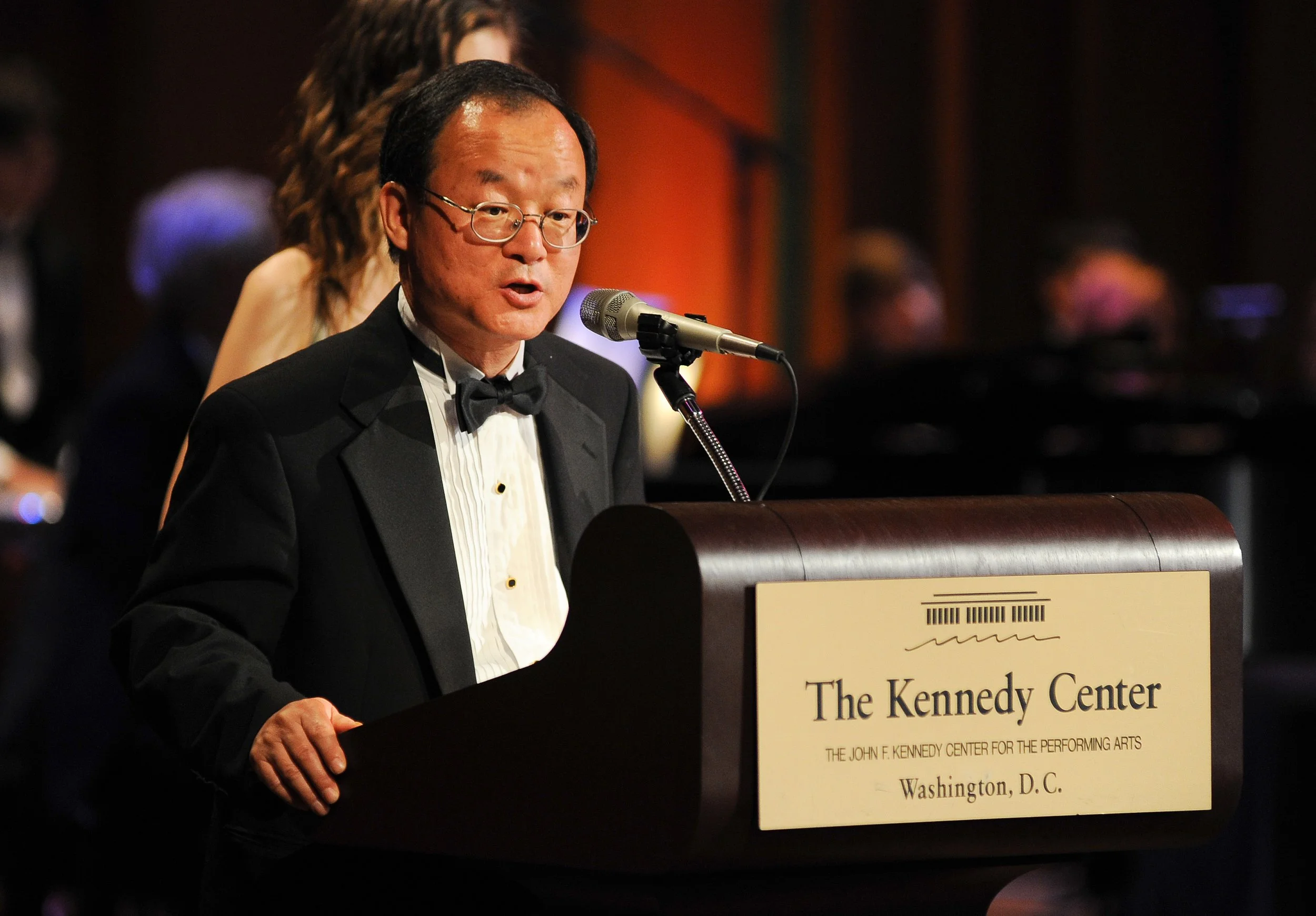 A man in a tuxedo and glasses speaking at a lectern at The Kennedy Center in Washington, D.C., with a microphone in front of him. The background is blurred with people behind him.