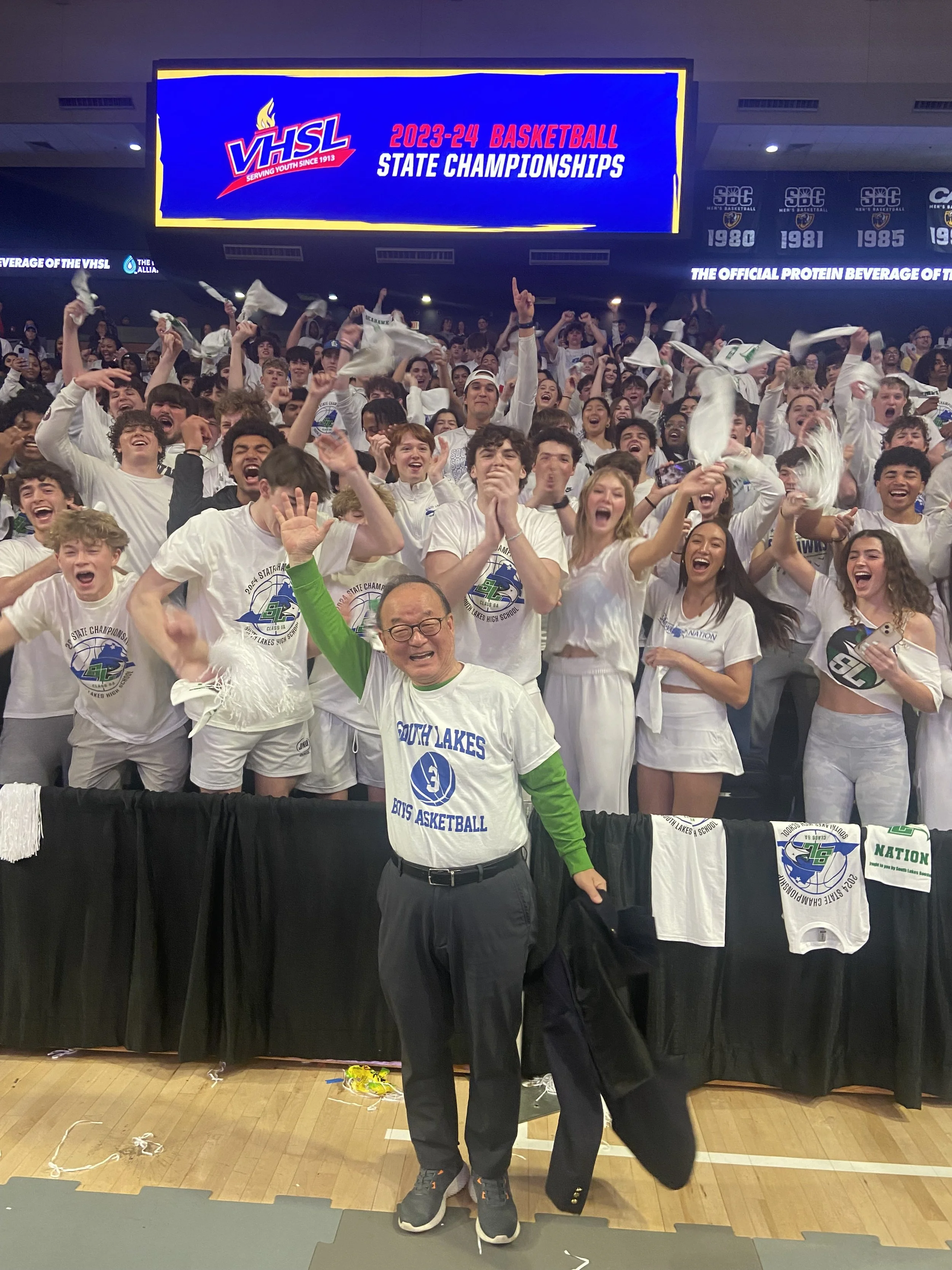 A large group of celebrating students and a man in front, in a gymnasium with a scoreboard and digital display. The students are wearing white t-shirts with a school logo, some are holding white towels, and there is a banner on the digital display th