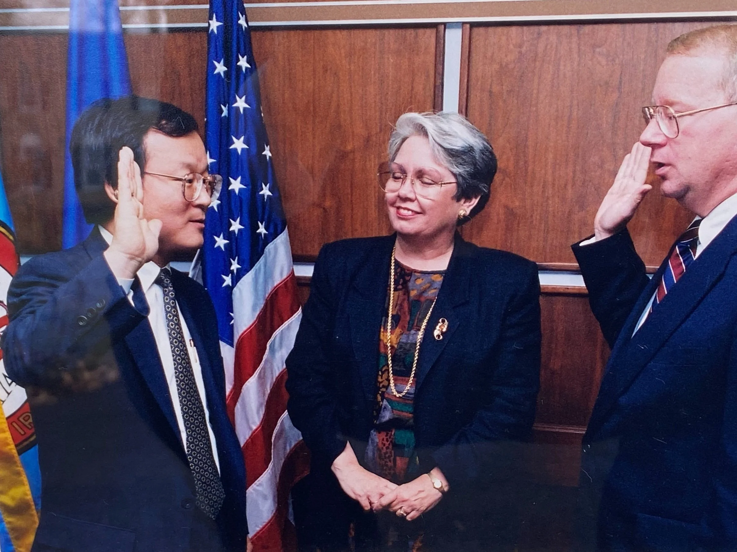 A man in a suit and glasses taking an oath while raising his right hand, a woman with short gray hair standing with her hands clasped in front of her, and another man in a suit and glasses raising his right hand in front of two flags, one of which is