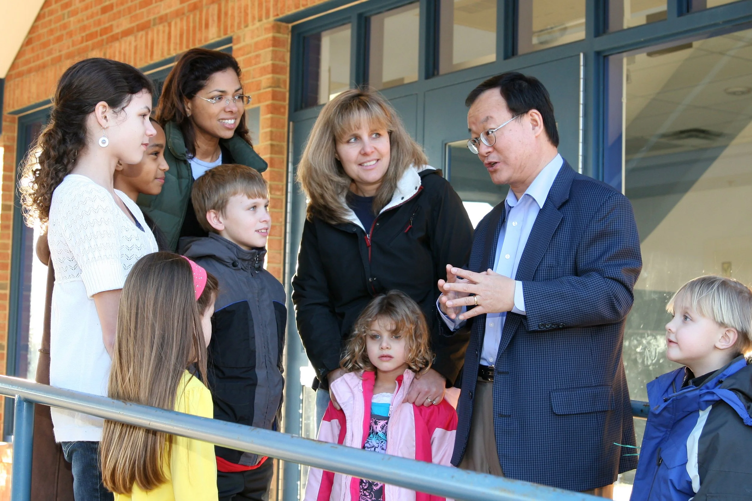 A man in a suit is speaking to a group of children and two women outside a building with brick walls and large glass windows.