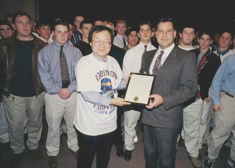 A man in a suit receives a framed certificate from another man wearing a Robinettes football sweatshirt, surrounded by a group of young men in casual and semi-formal attire.