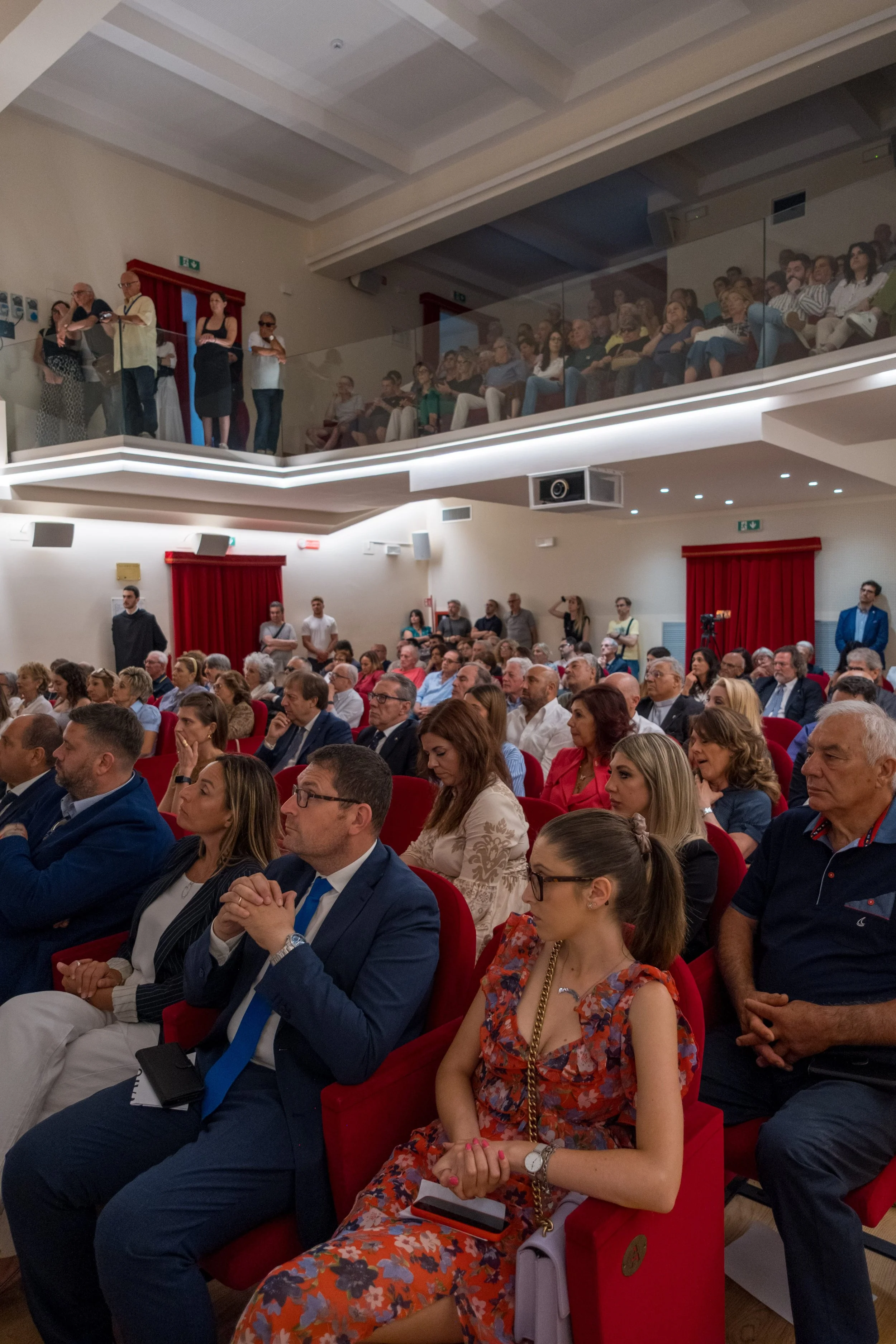 Un pubblico seduto in un auditorium durante un evento, con un secondo livello di persone che si trova in balconata.