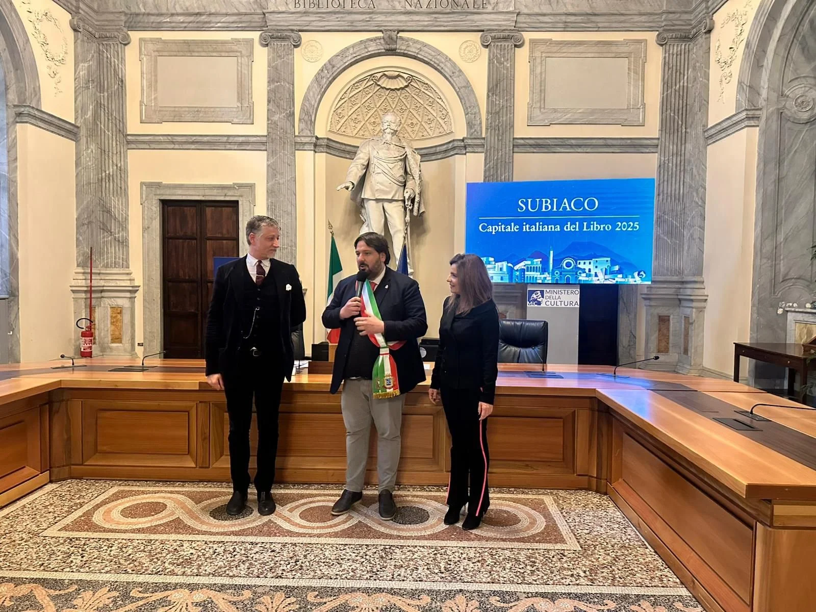 Tre persone sono in piedi in una stanza decorata con dettagli classici e una statua al centro della parete dietro di loro. Sullo sfondo, uno schermo blu mostra le parole "SUBIACO Capitale italiana del Libro 2025" e il logo del Ministero della Cultura