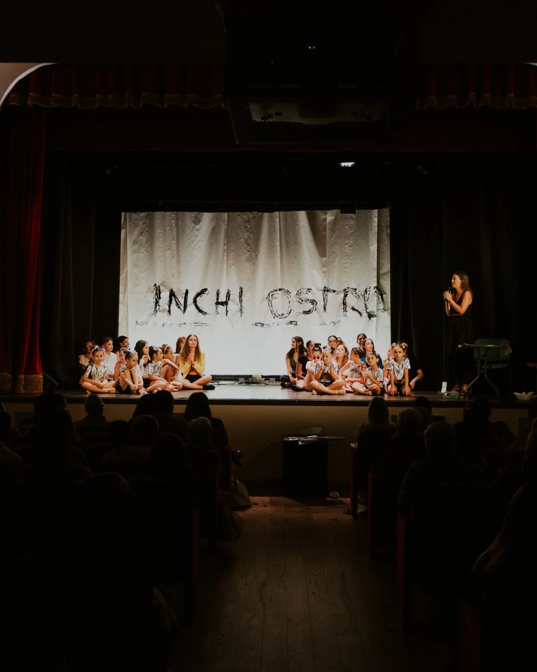 Un gruppo di bambini seduti sul palco durante uno spettacolo teatrale, con un'adulta che si trova sul lato destro che parla al pubblico, con uno sfondo con la scritta 'INCHIOSTRA'