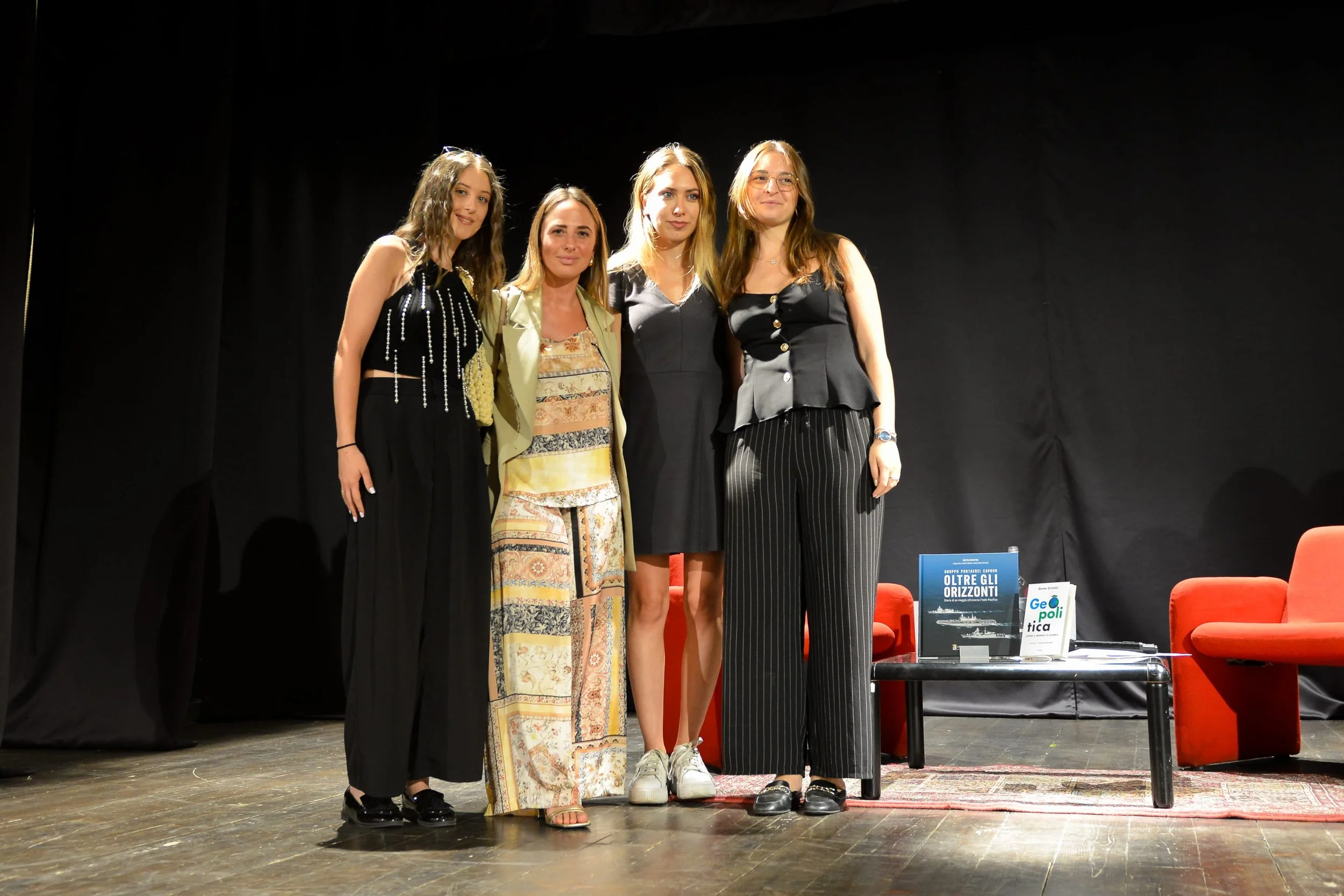 Quattro donne in piedi su un palco, posano per una foto davanti a uno sfondo nero, con alcuni mobili rossi visibili a destra.