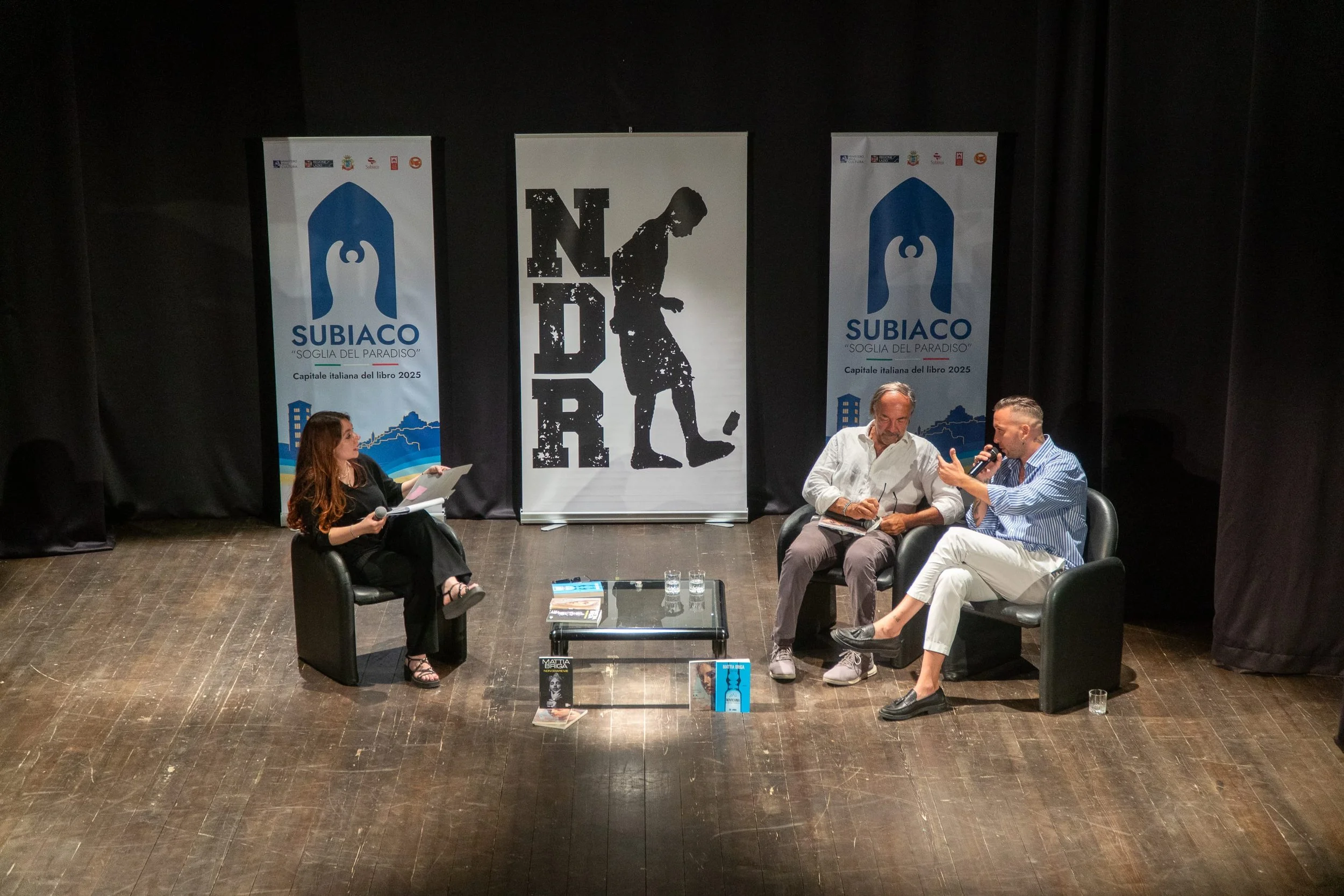 Tre persone sedute su un palco durante un evento letterario. Due uomini parlano tra loro e una donna ascolta con un libro in mano. Sullo sfondo, pannelli con il logo di Subiaco e un disegno stilizzato di una figura umana.