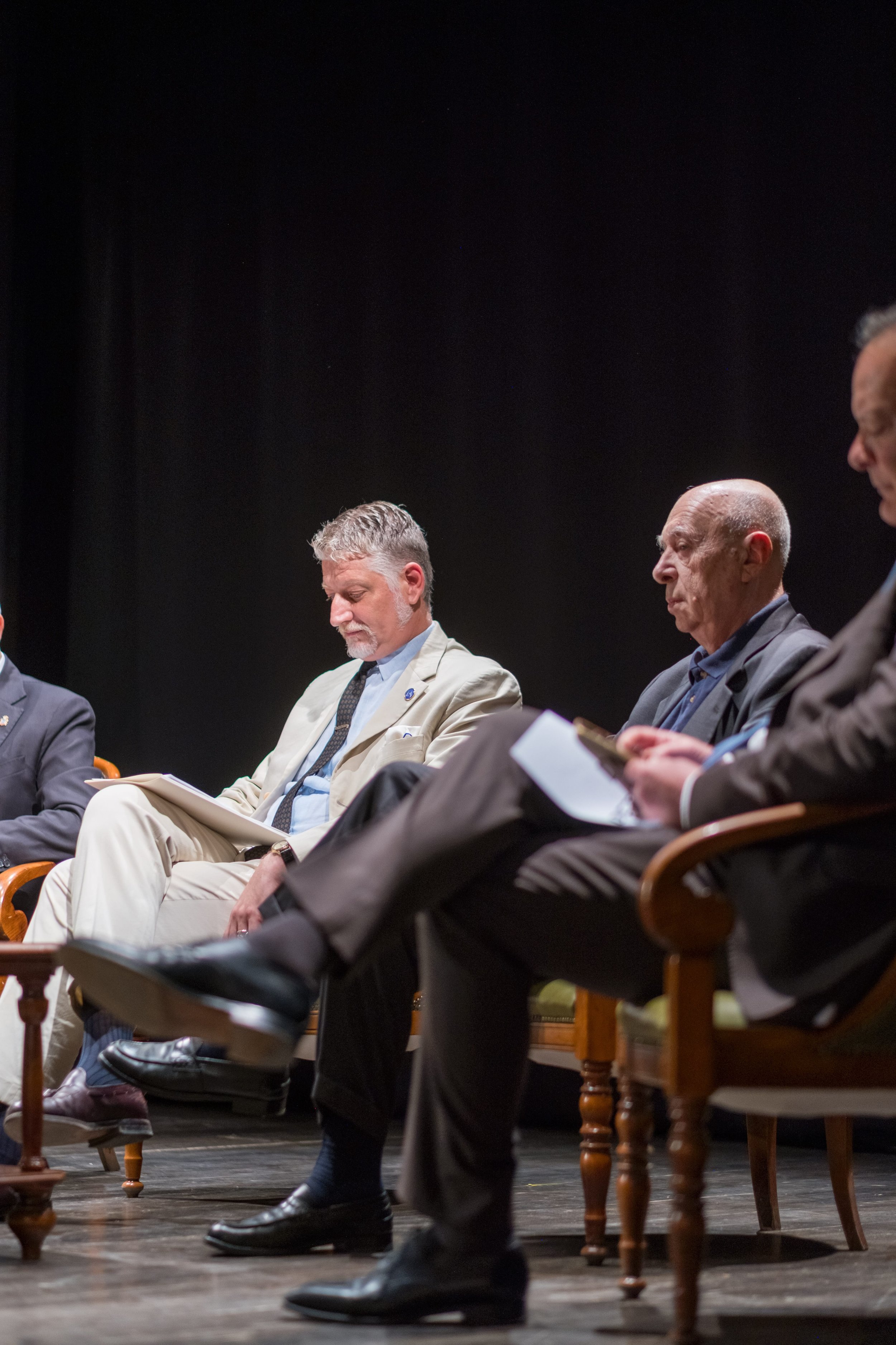 Azienda in giacca e cravatta, in discussione, seduti su sedie di legno, con altri uomini in abiti formali, in un ambiente di conferenza o dibattito.
