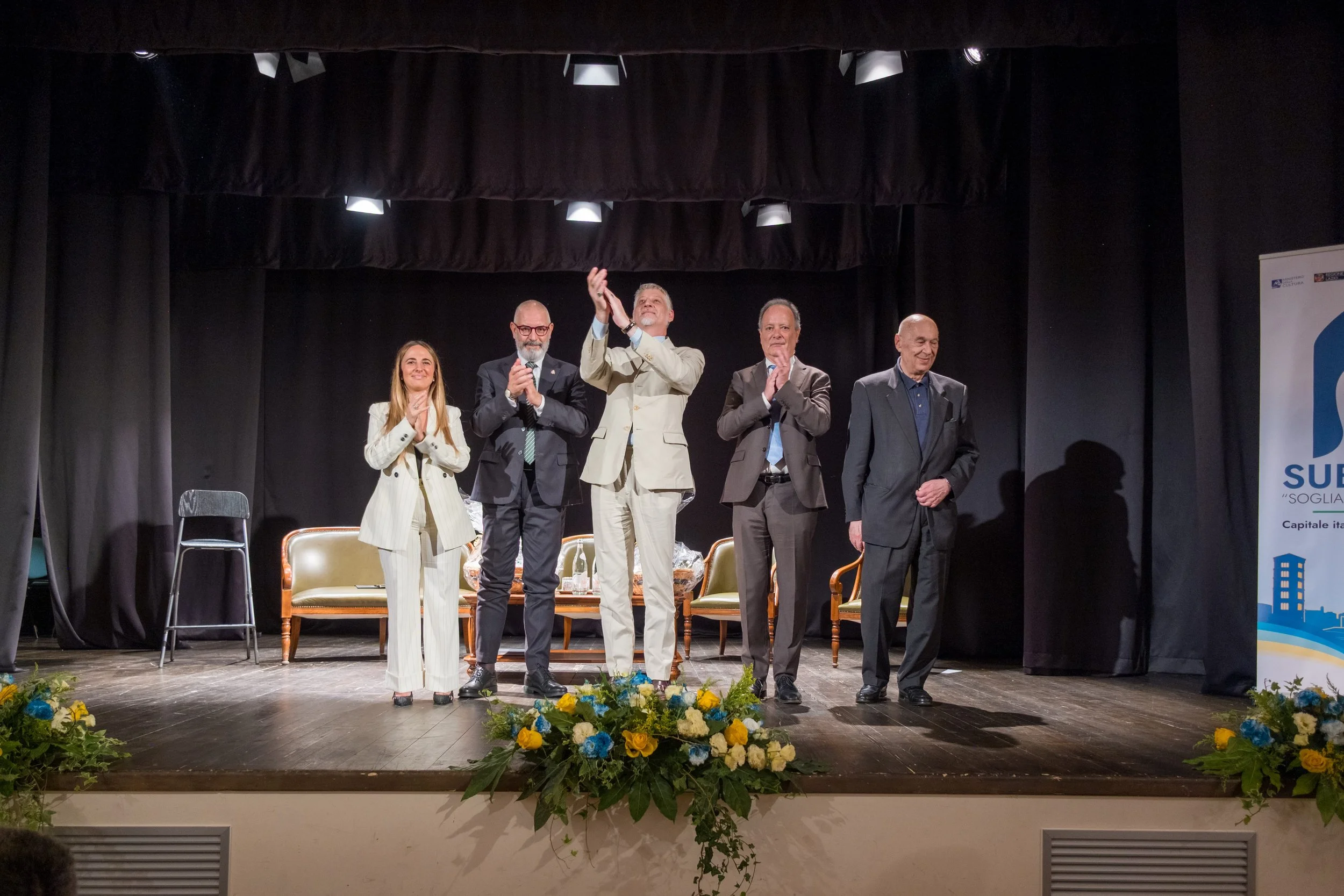 Cinque persone sul palco di una conferenza o evento, applaudendo, con decorazioni floreali in primo piano, e uno schermo con loghi e testo sul lato destro.