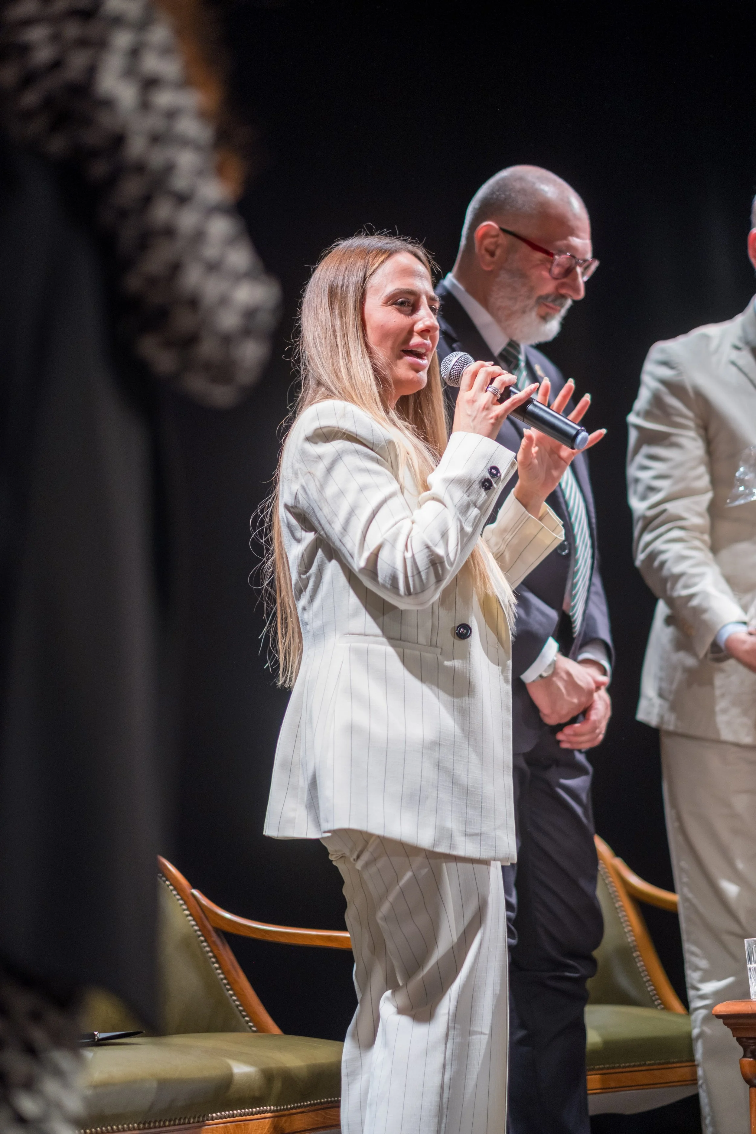 Donna con capello lungo e vestito a righe bianche e beige parla con microfono seduta su una poltrona d'epoca in un evento pubblico.