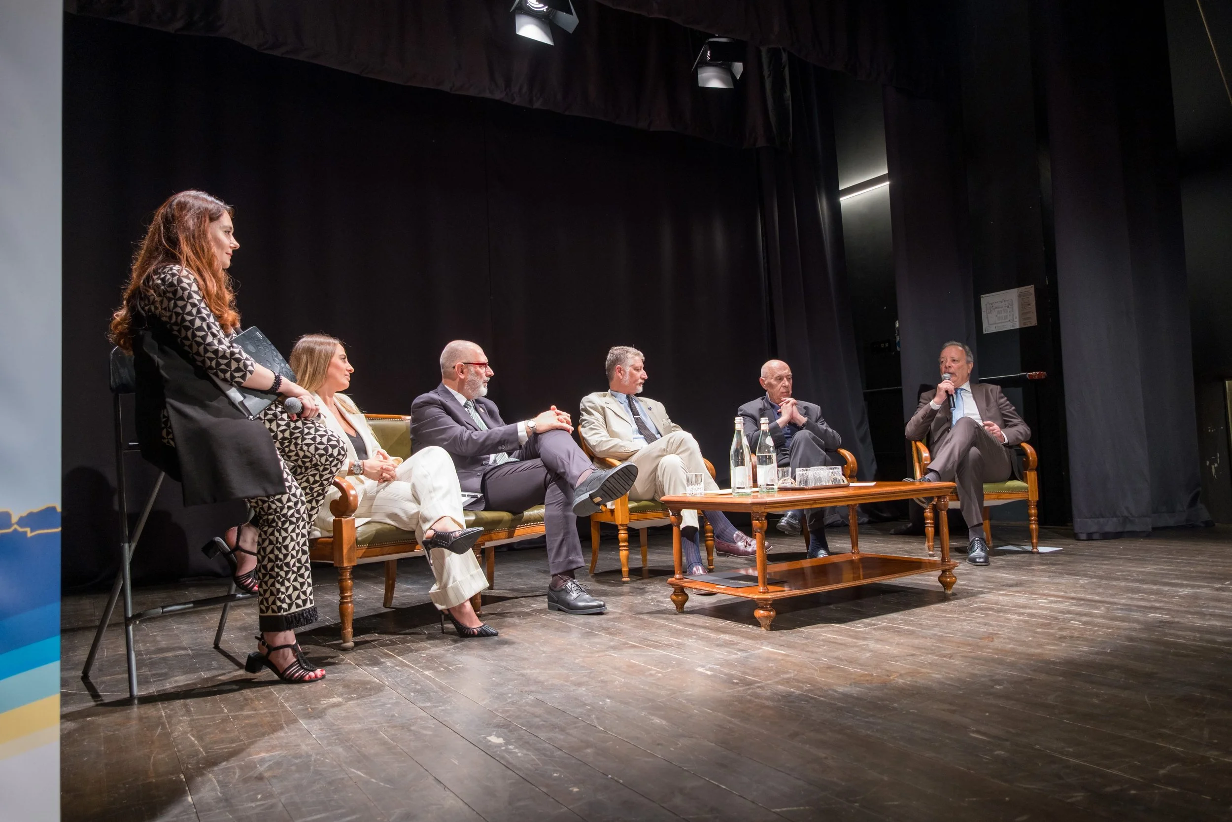 Panel di sette persone sedute e una donna in piedi con microfono in un evento su un palco con sfondo nero.