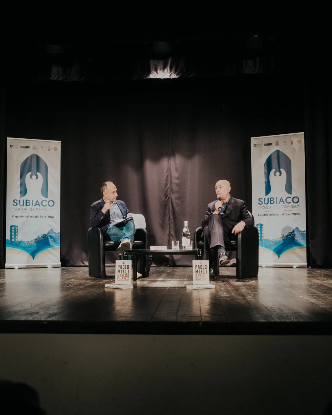 Due uomini seduti su poltrone nere durante un confronto o intervista sul palco, con un tavolino tra loro che ha due libri e una bottiglia d'acqua. Due banner pubblicitari con il logo di Subiaco e il titolo 'Soglia del Paradiso' sono sullo sfondo.
