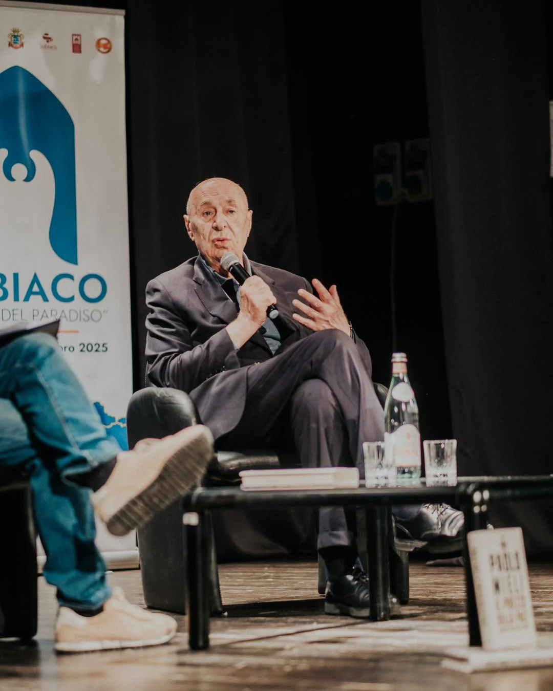 Un uomo anziano seduto che parla in un microfono durante un evento, con uno sfondo con un logo e testo in italiano.