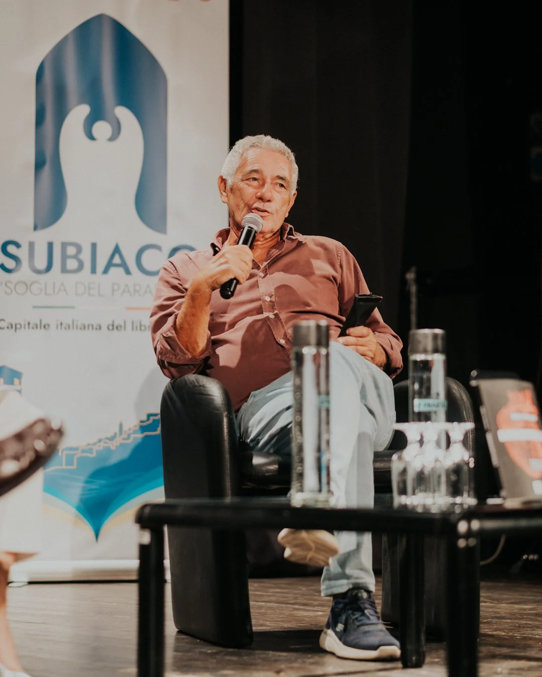 Un uomo anziano con capelli grigi, vestito con una camicia marrone e pantaloni chiari, seduto su una sedia di fronte a un pubblico, tiene in mano un microfono e uno smartphone. Sullo sfondo, un banner con il logo di SUBIAC e la scritta 'Capitale ital