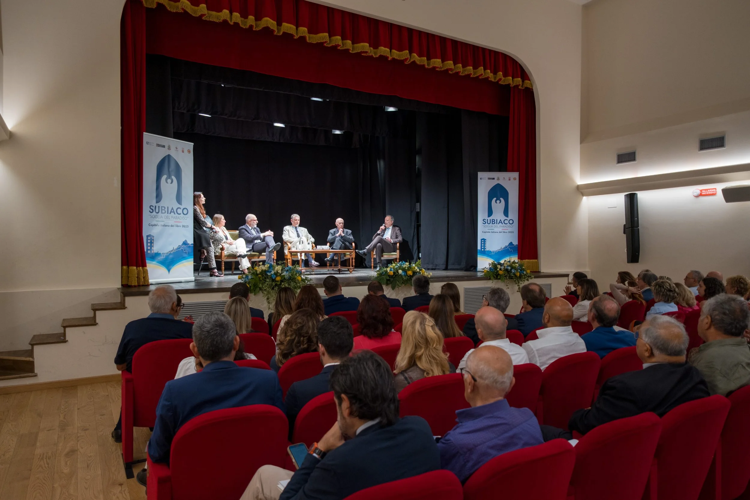 Un dibattito o conferenza su un palco con sei persone sedute in piedi e una che parla, con il pubblico seduto davanti. Ci sono due cartelloni con il logo 'SUBIACO'.