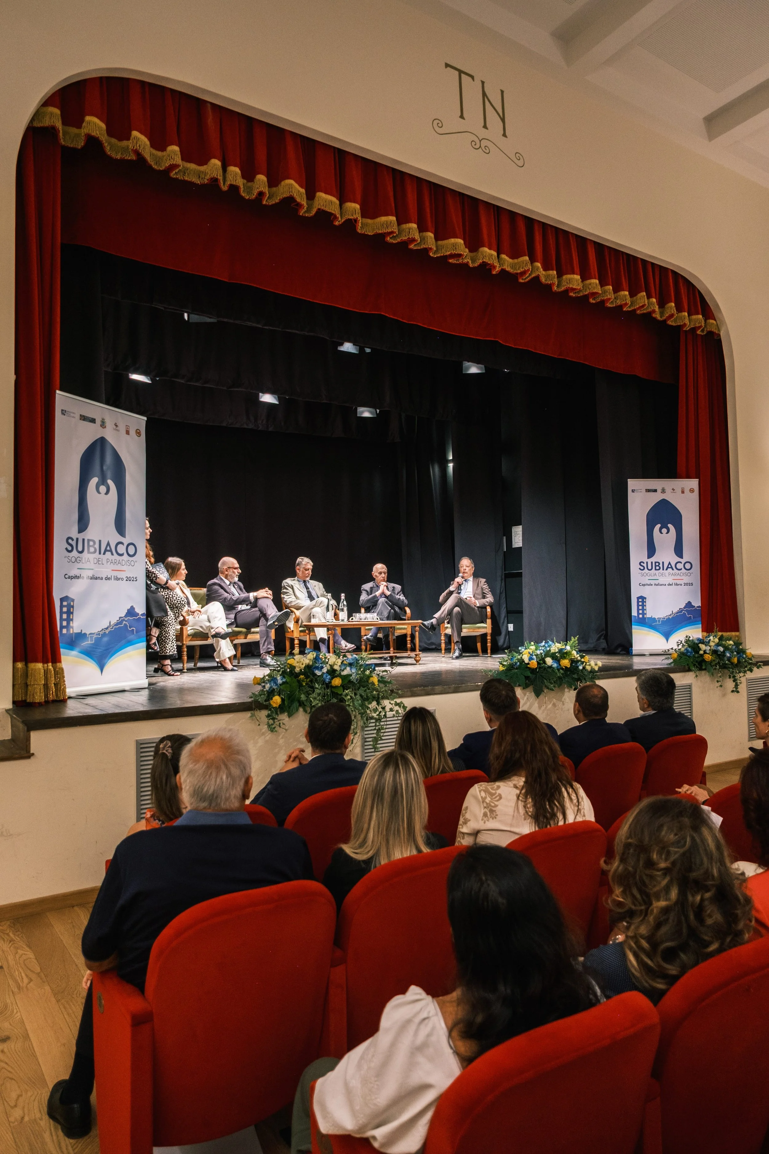 A conference or panel discussion with multiple speakers on stage, seated on chairs, with banners displaying 'SUBIACO' and 'SOGNE DEL PARADISO', and an audience seated in red chairs watching the event.