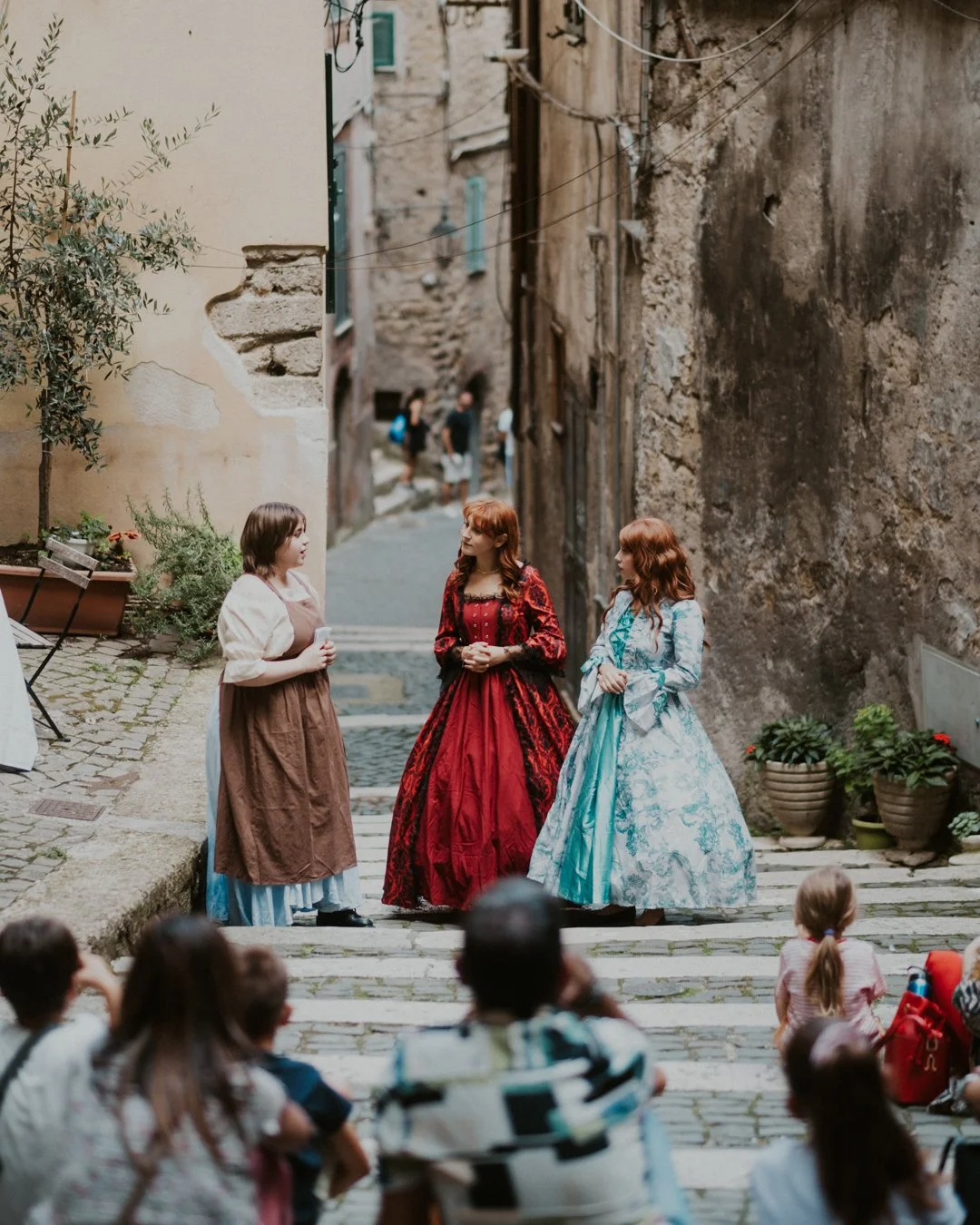 Tre donne vestite con abiti storici, che si confrontano in un vicolo di pietra, con un pubblico di bambini e adulti seduti di fronte, in un ambiente che ricorda un borgo antico.