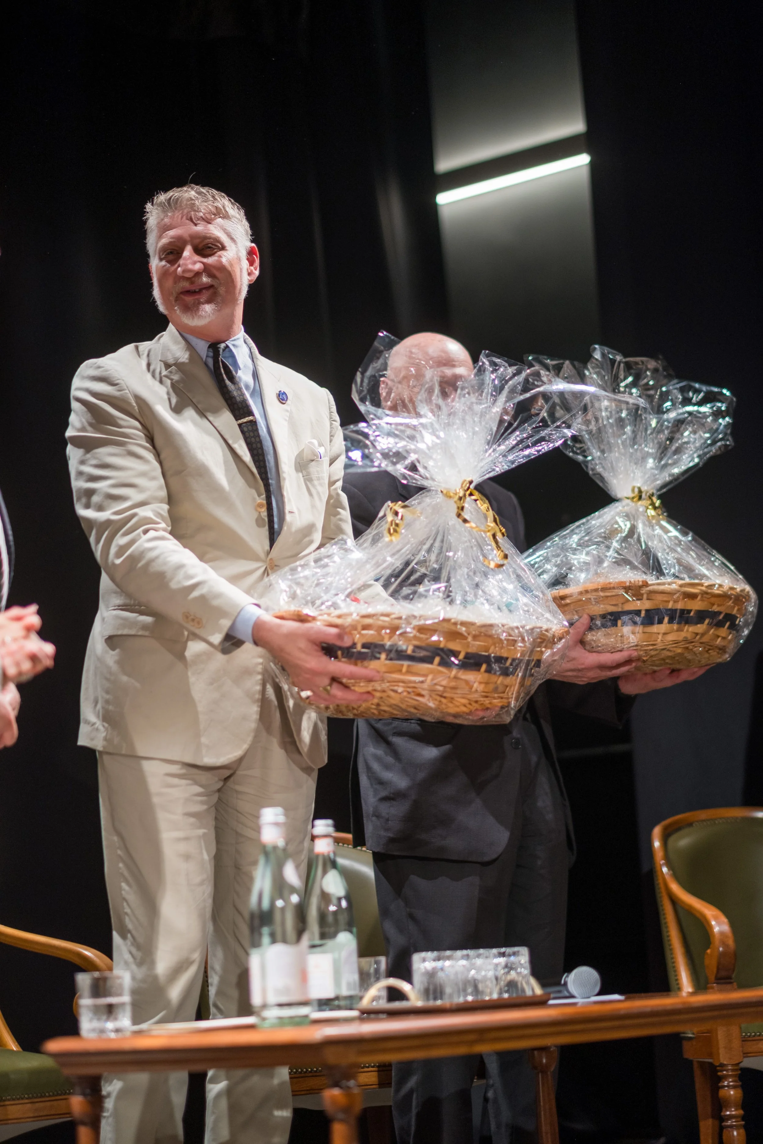 Due uomini sorridenti presentano dei cesti regalo avvolti in pellicola trasparente durante un evento, con tavolo e bottiglie d'acqua sullo sfondo.