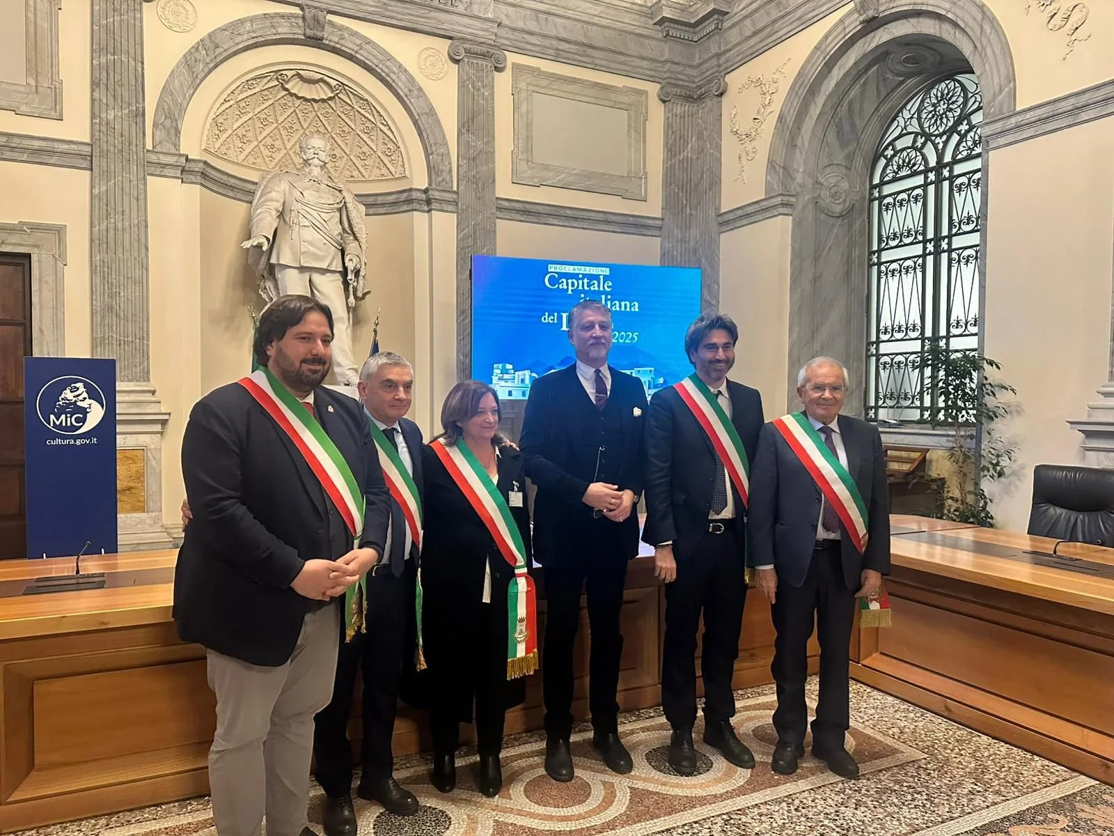 Sette persone in abiti formali con fascia tricolore, in piedi in una sala elegante con statue e finestre con grate, durante un evento ufficiale in Italia.