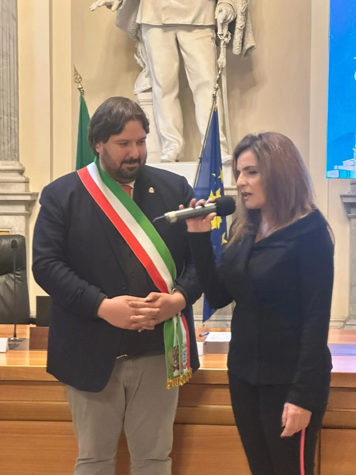 Un uomo con fascia tricolore viene intervistato da una donna in un ufficio con bandiere europee e italiane sullo sfondo.