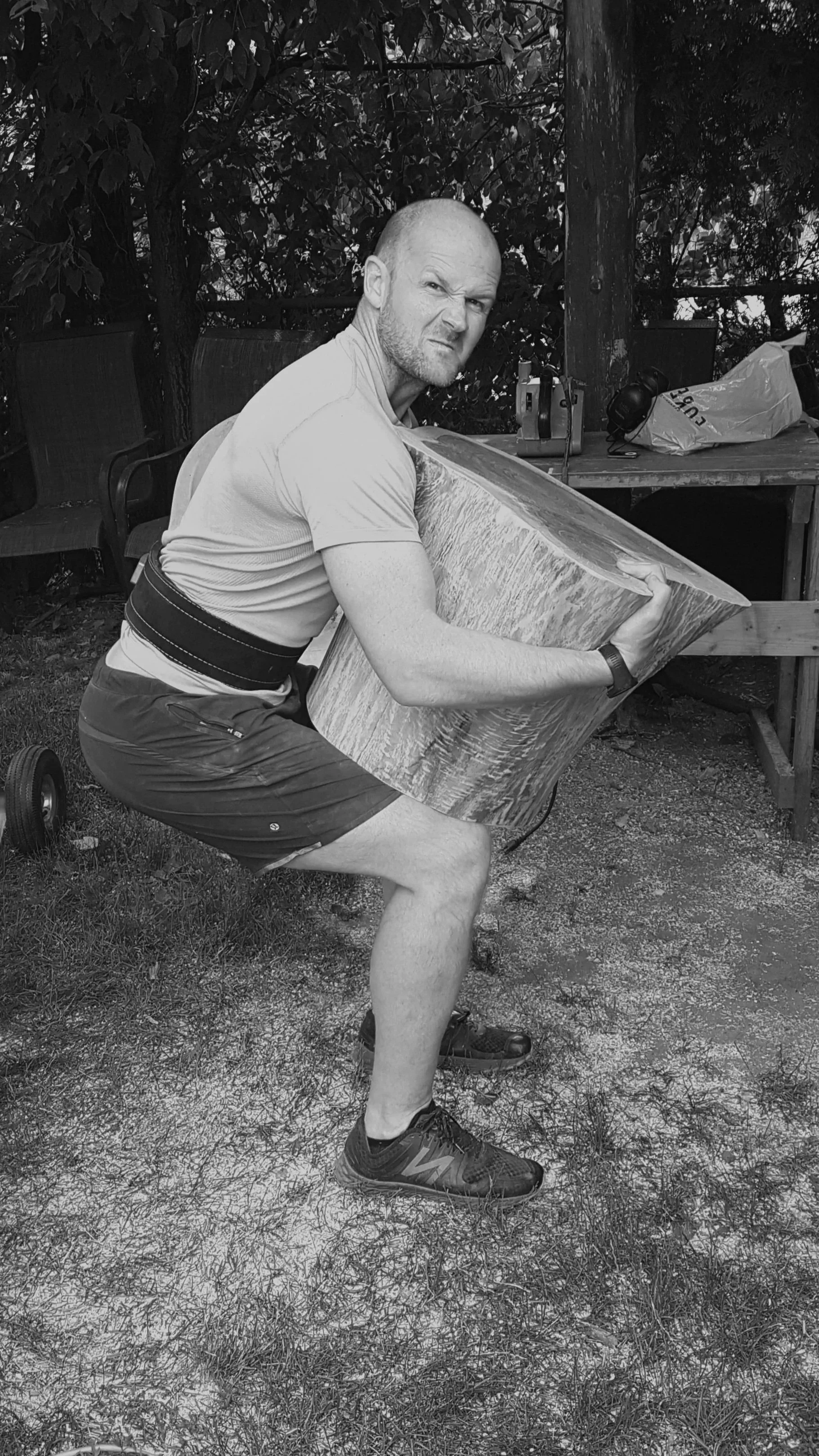 Gavin Peacock wearing a weightlifting belt is crouched down outdoors, hugging a large, round, walnut stump side table with a serious facial expression.