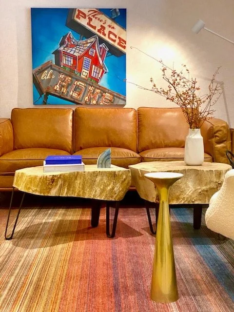 Living room with a brown leather sofa, a colorful abstract painting that says 'Welcome to the Place' hanging on the wall behind the sofa, a white vase with dried branches on a side table, a rustic wooden coffee table with books and a decorative object, a striped multicolored rug, and a white floor lamp.