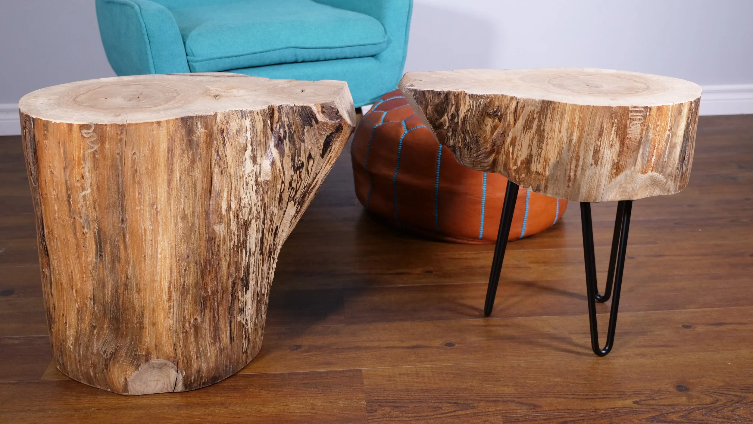 Two wooden Oak side tables made from tree trunks, one large and one small, with black metal hairpin legs in a living room with hardwood floor, light blue armchair, brown leather pouf, and a white wall.