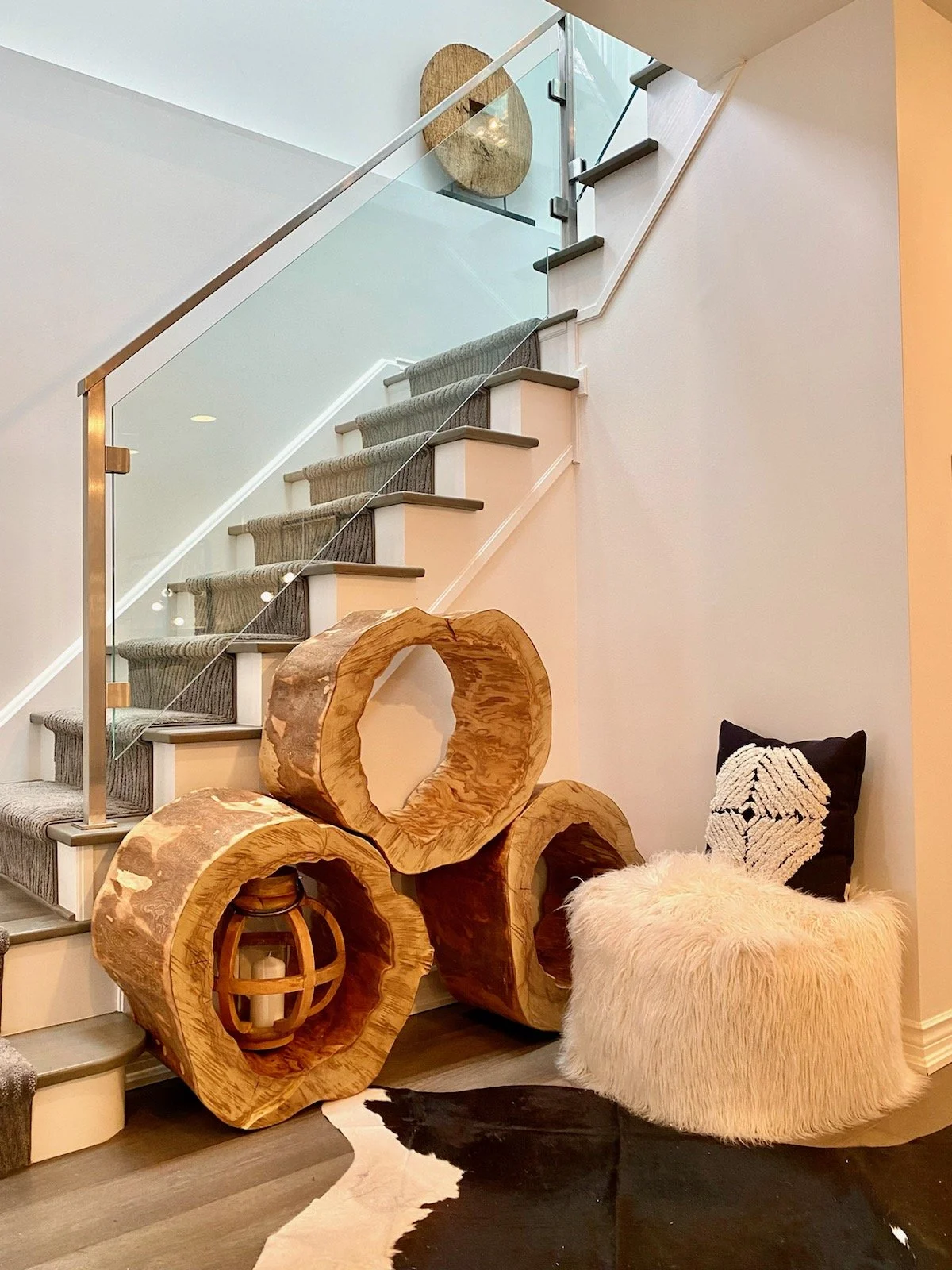 Interior view of a staircase with glass railing, modern wooden log sculptures, a beige furry chair, and decorative pillows. The staircase has dark treads and a light-colored wall, with a piece of abstract wall art visible at the top.