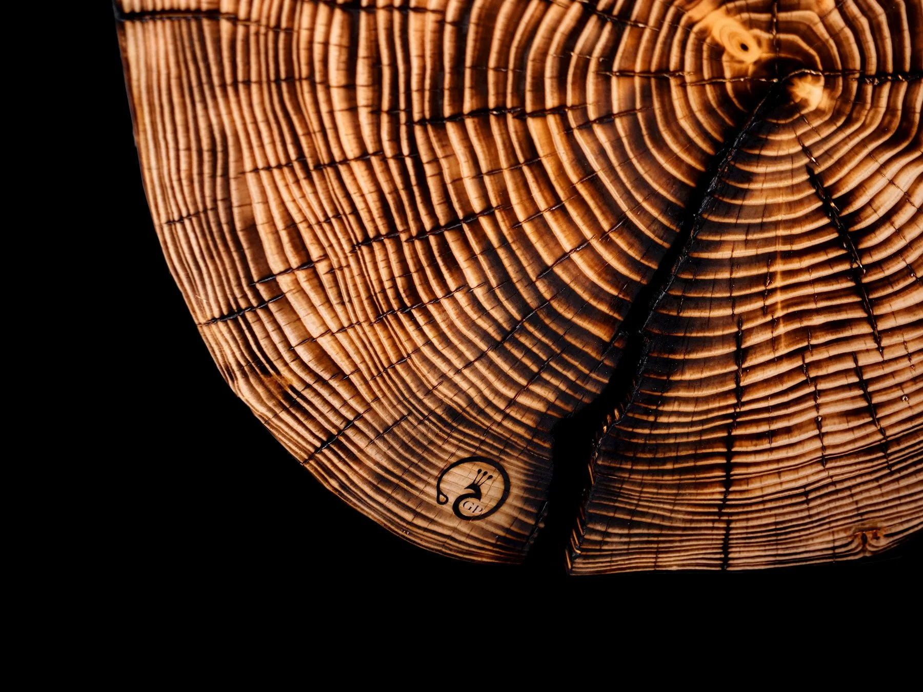 Japanese Shou Sugi Ban wood burning technique Close-up of a wooden tree trunk cross-section with growth rings, a small circular maker's mark logo near the bottom left, and a crack running through the rings.
