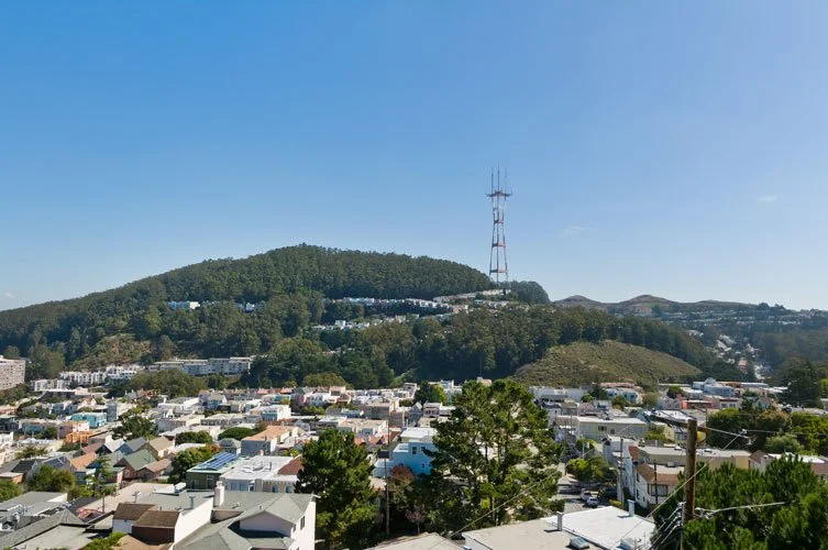 View of a town with closely packed houses, a hill covered in trees, and a tall communication tower on top of the hill under a clear blue sky.