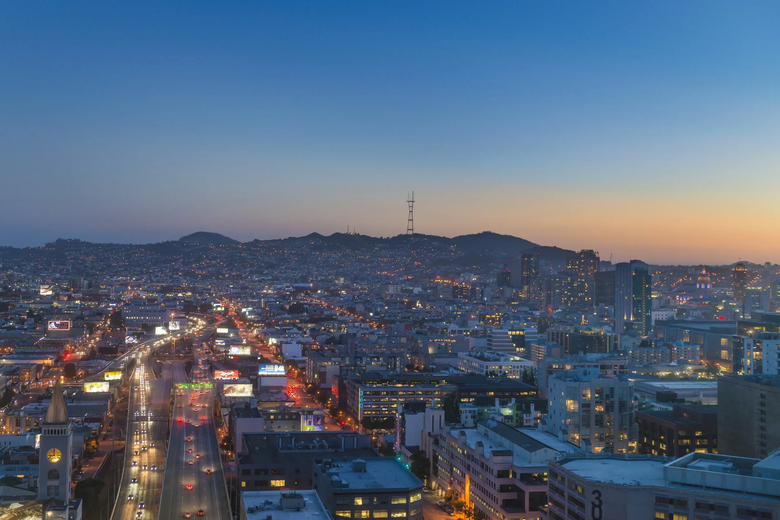 City skyline at dusk with illuminated buildings, busy roads, and a hill with a tower in the background