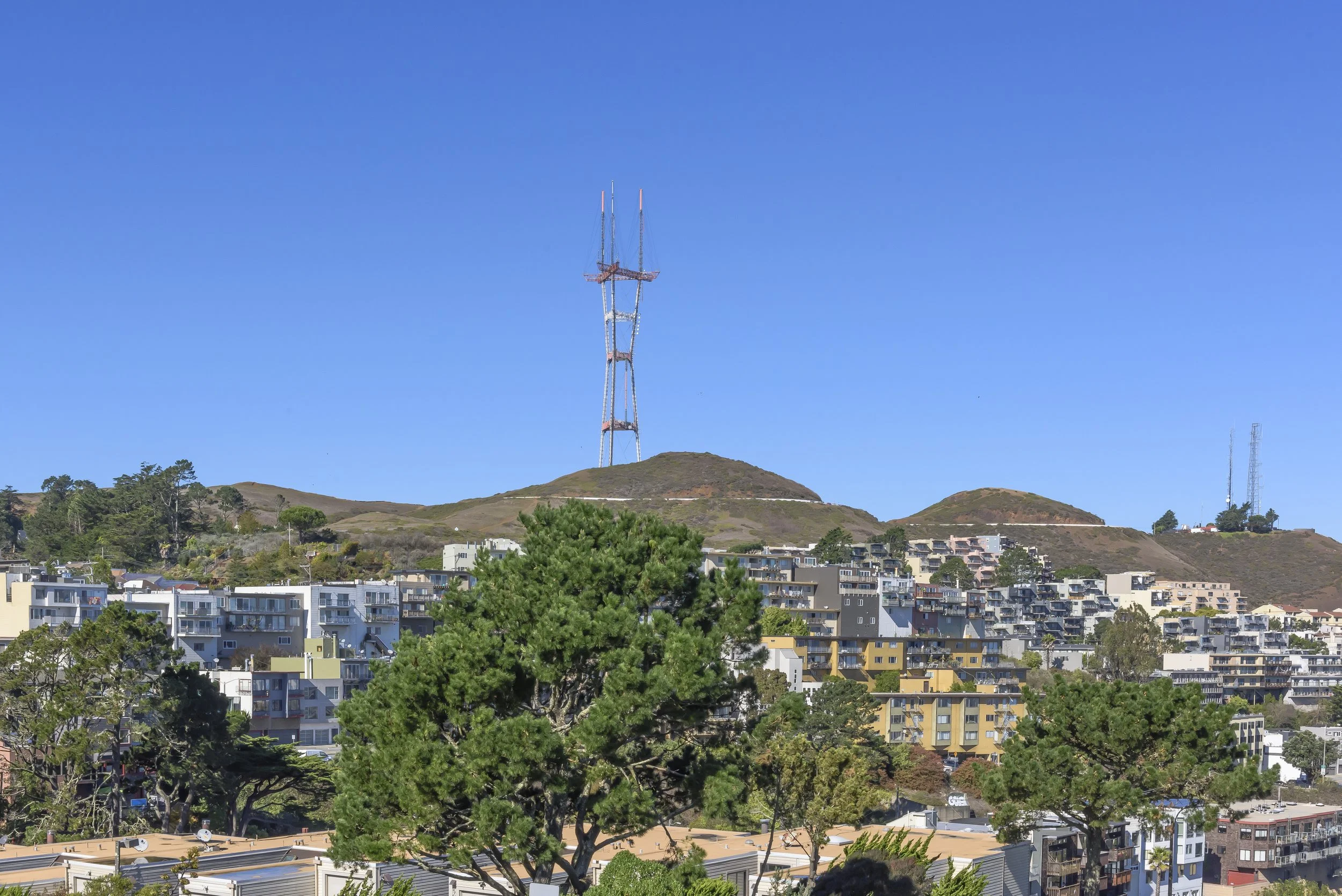 Cityscape with hills, trees, and buildings beneath a clear blue sky, with a large radio tower on a hilltop.