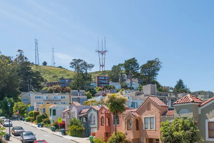 Colorful houses line a street, with hills and trees in the background. Tall communication towers are visible on the hilltop under a clear blue sky.