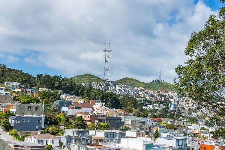 View of a hilly residential area with colorful houses, green trees, and a tall radio tower in the background under a partly cloudy sky.