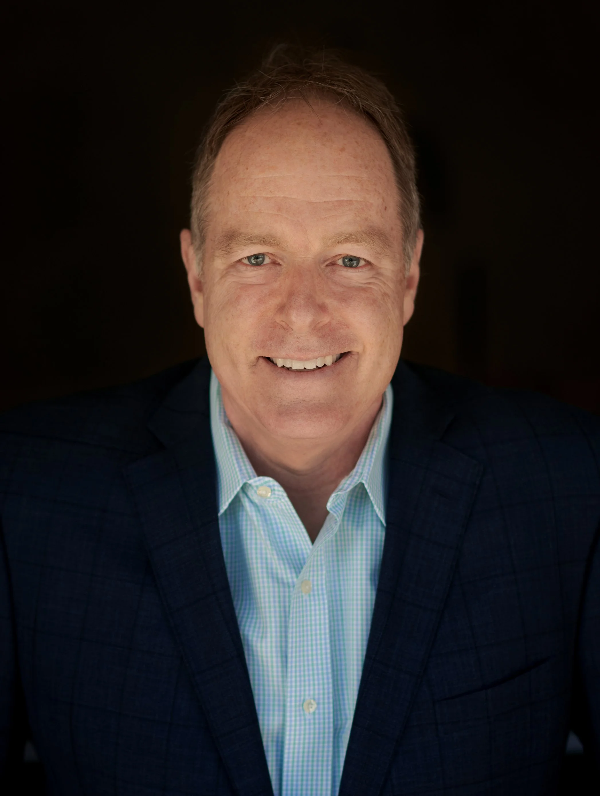 Close-up portrait of a smiling middle-aged man with short light brown hair, blue eyes, wearing a blue checked shirt and a dark blazer.