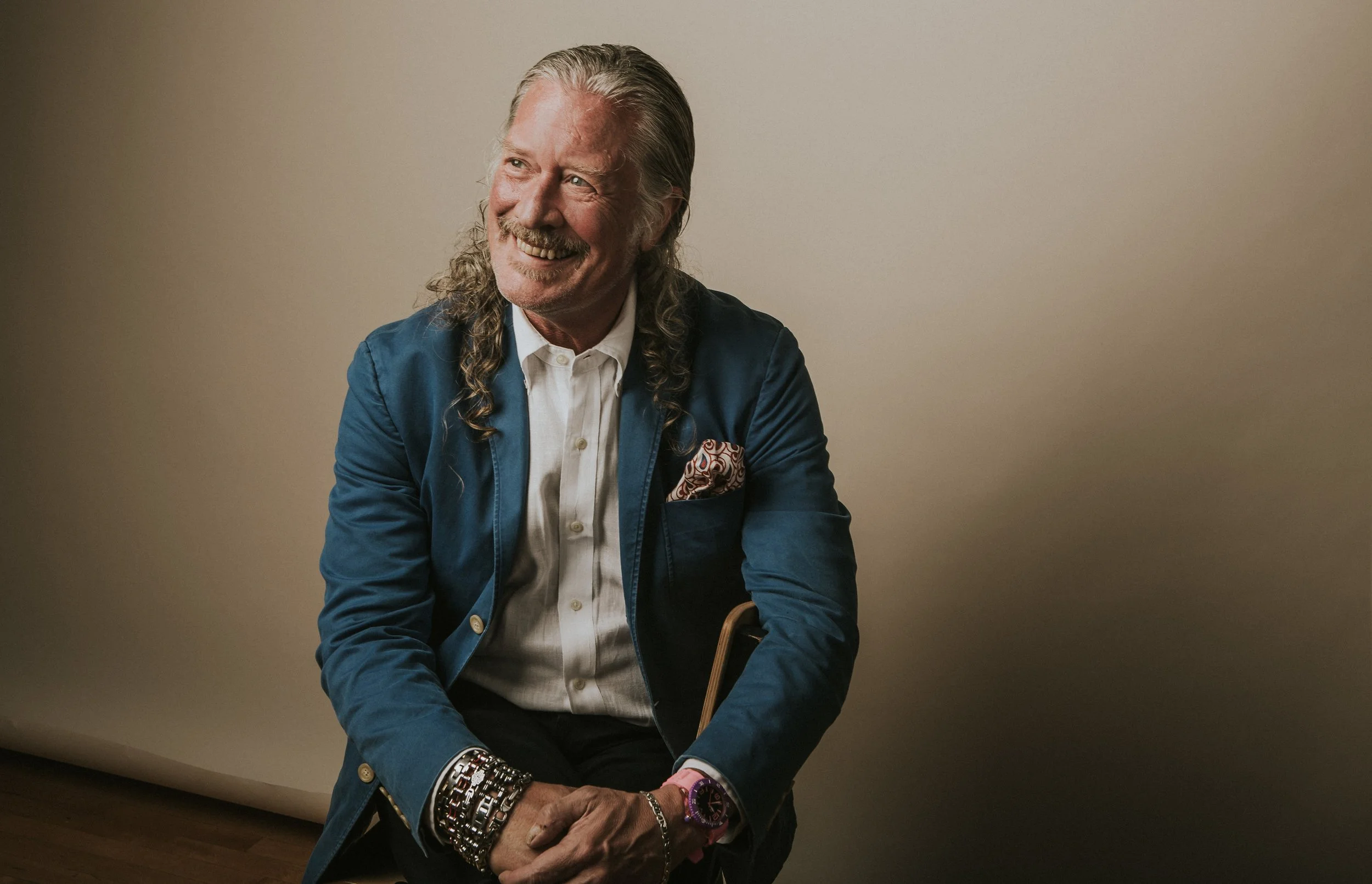 A smiling man with long gray hair and a beard, wearing a blue blazer with a paisley pocket square, white dress shirt, and multiple bracelets and a watch, sitting against a plain beige background.