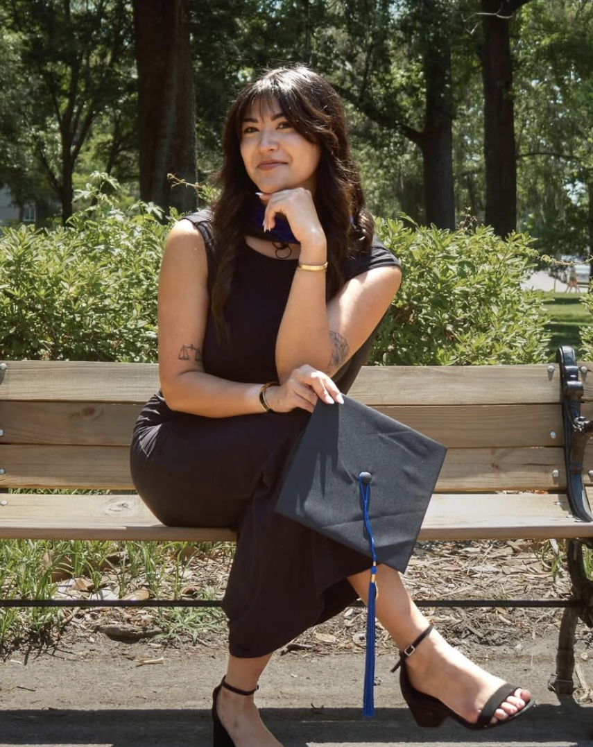 A woman in a black dress sitting on a park bench holding a graduation cap, with trees and greenery in the background.