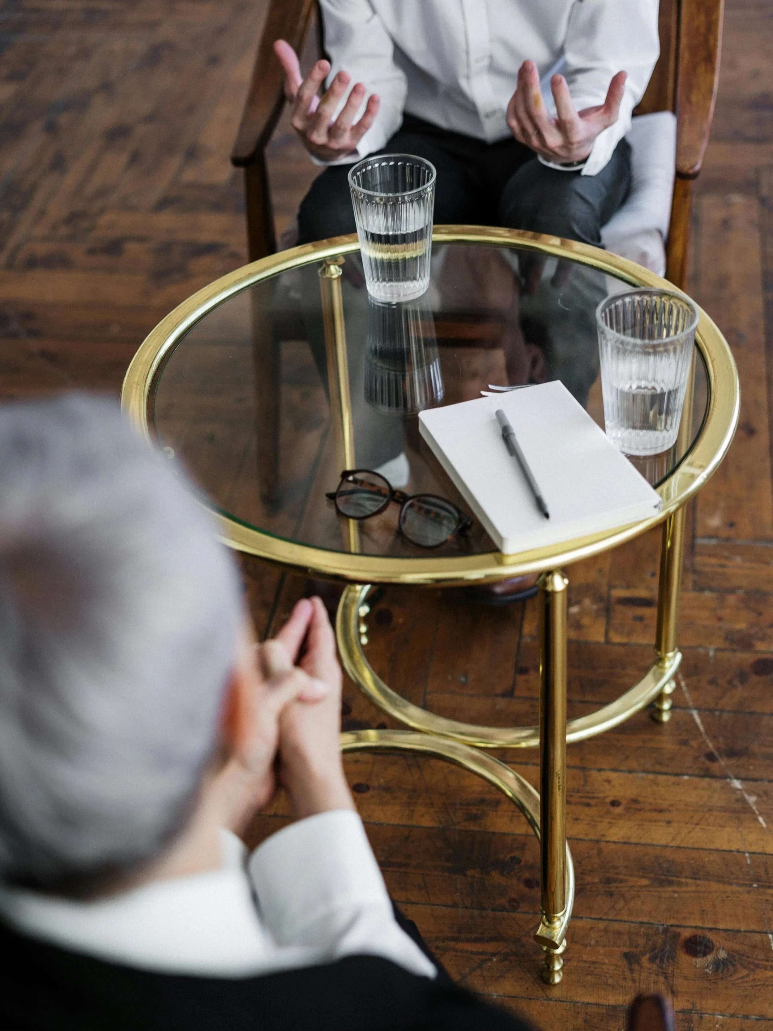 An interview or meeting scene with two individuals, one with gray hair and the other with dark hair, seated at a glass-top table with gold trim. On the table are two glasses of water, a notepad, a pen, and a pair of glasses. The person with gray hair appears to be listening attentively, while the person with dark hair is speaking with hand gestures.