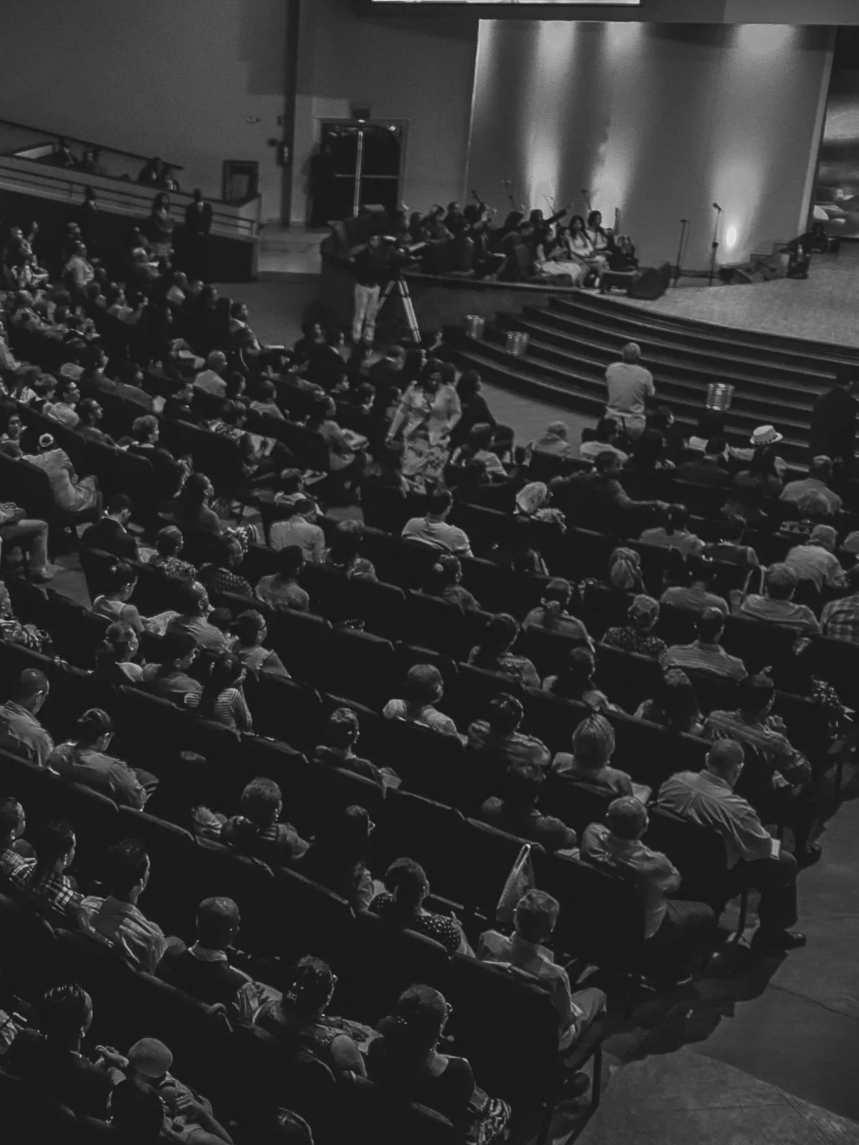 A large audience seated in a theater, facing a stage with a group of people and microphones, with some people standing near the stage.