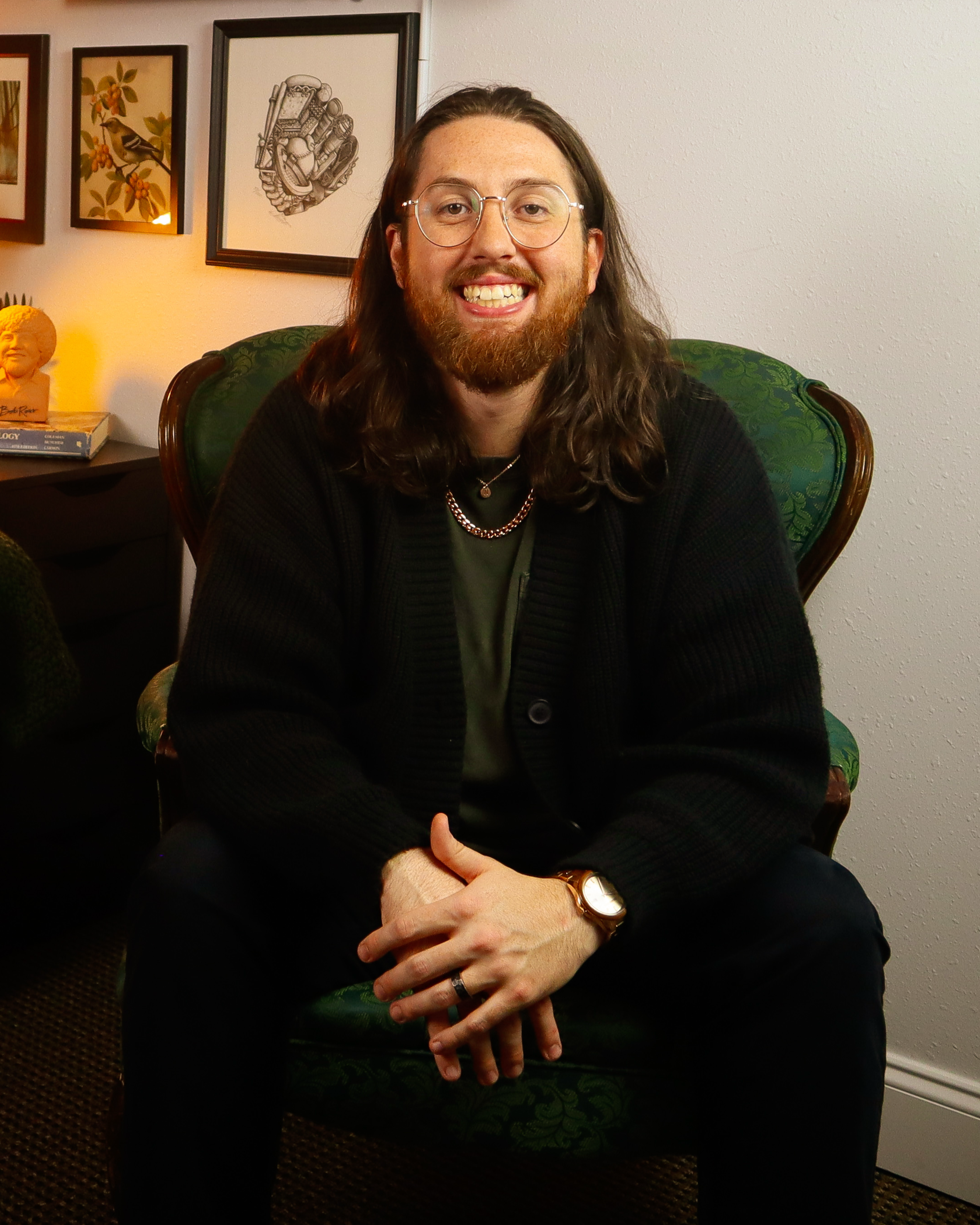 A smiling man with long hair and glasses sitting in a green armchair in a room with framed artwork on the wall behind him.