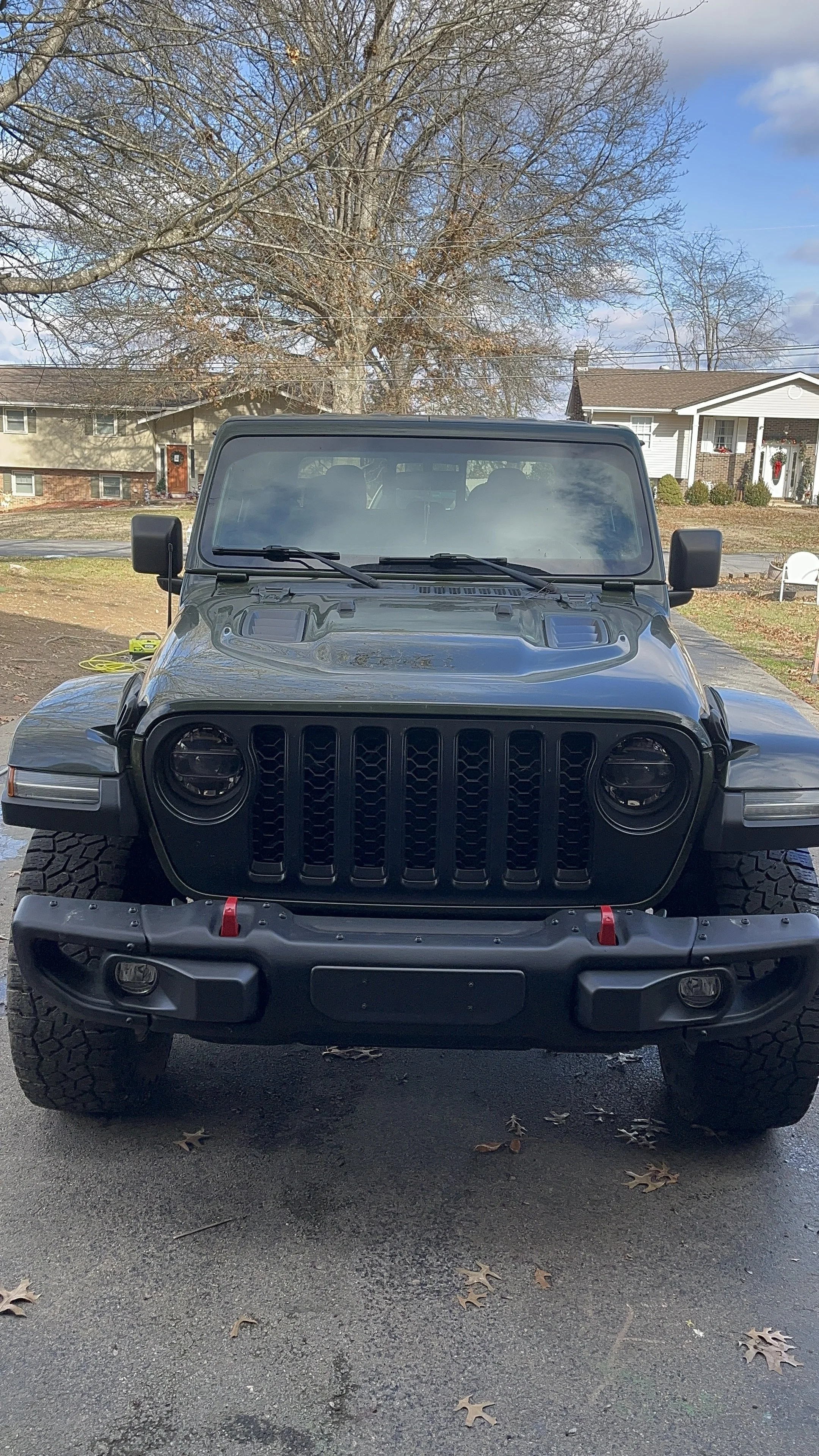 Front view of a black Jeep Wrangler parked on a driveway during daytime, with leafless trees and houses in the background.
