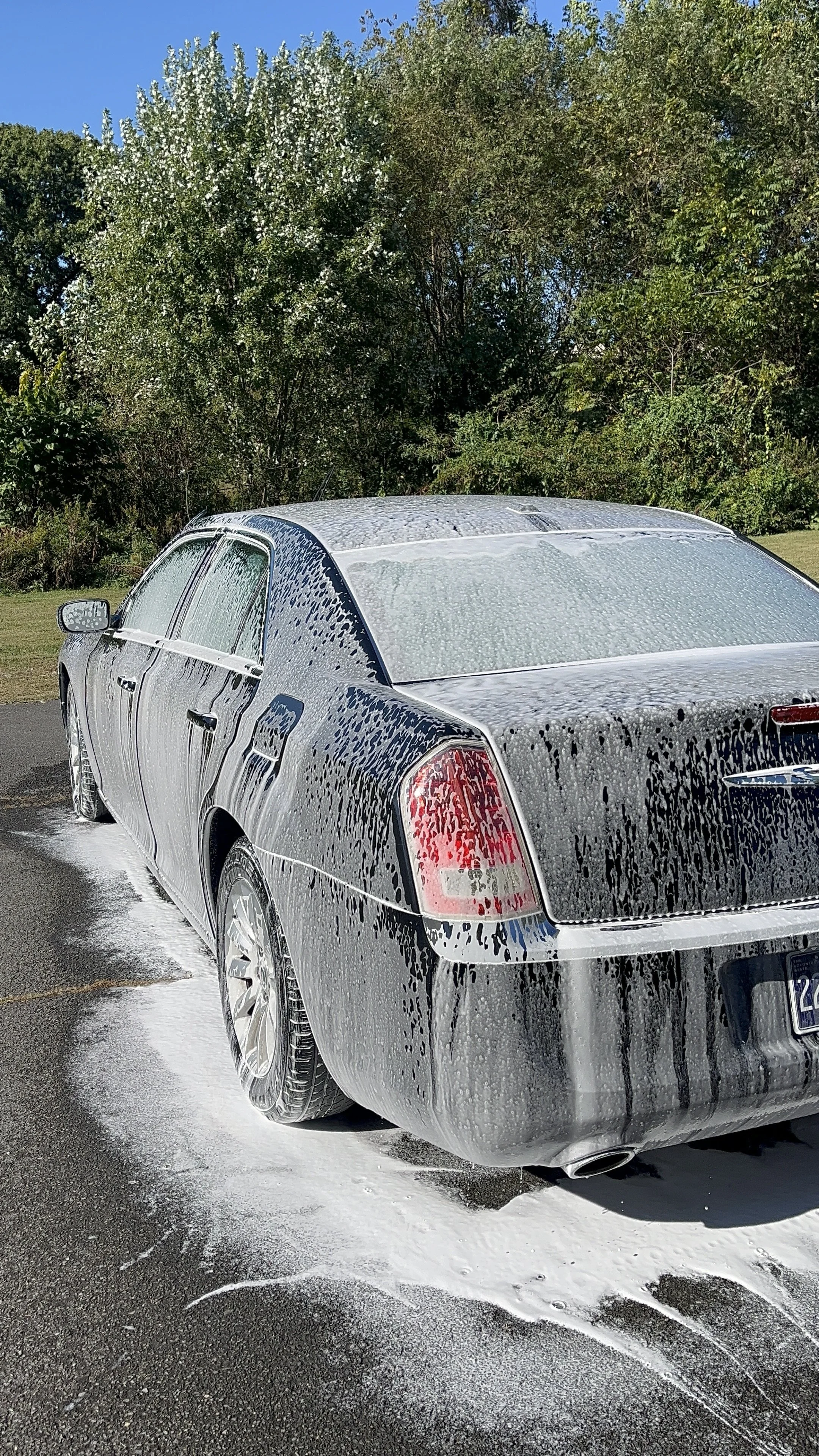 A black sedan car being washed with soap, creating foam on the car's surface, on a paved outdoor area with trees and blue sky in the background.