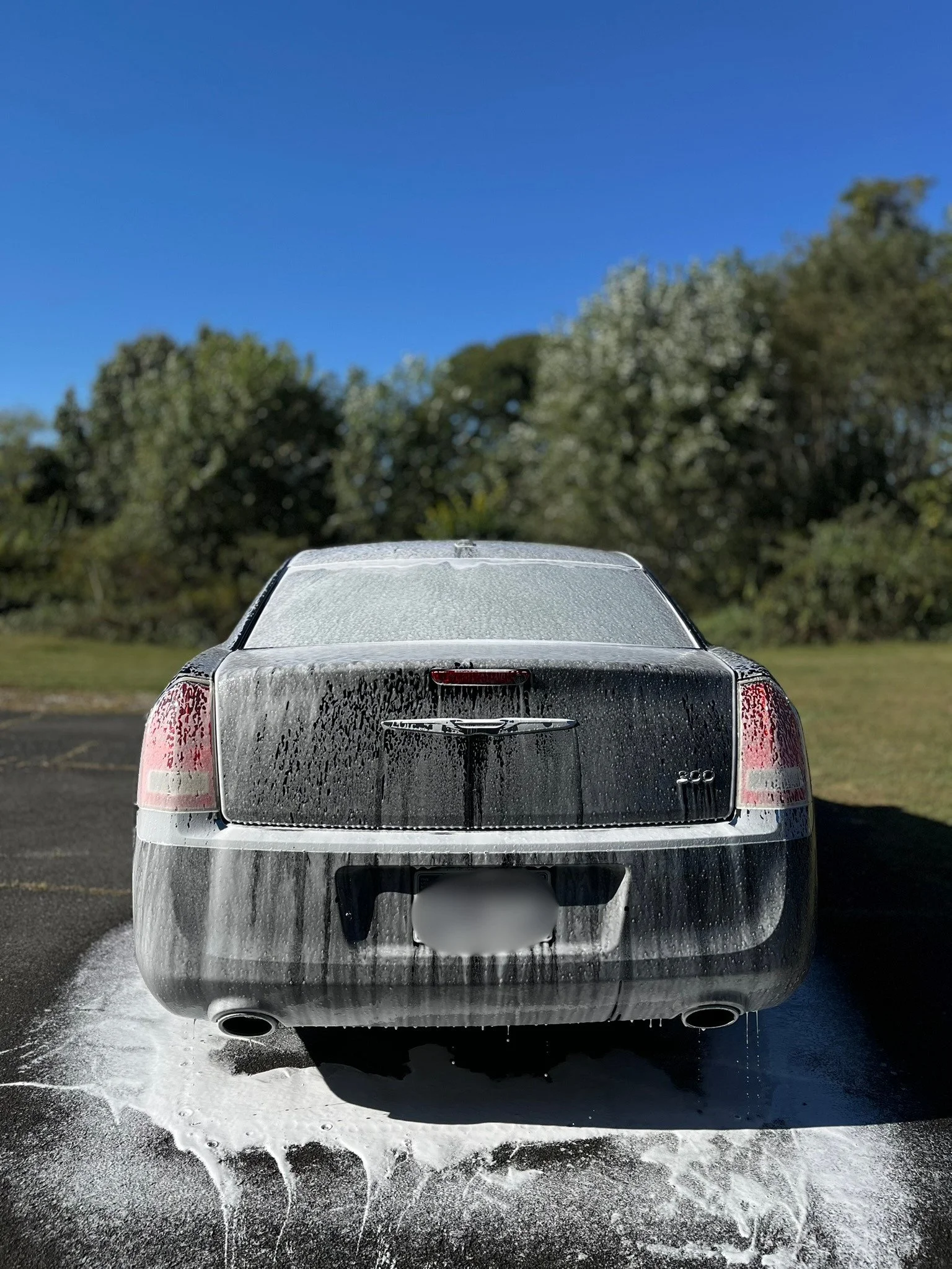 Back view of a car being washed with soap and water, on a parking lot near trees and a clear blue sky.