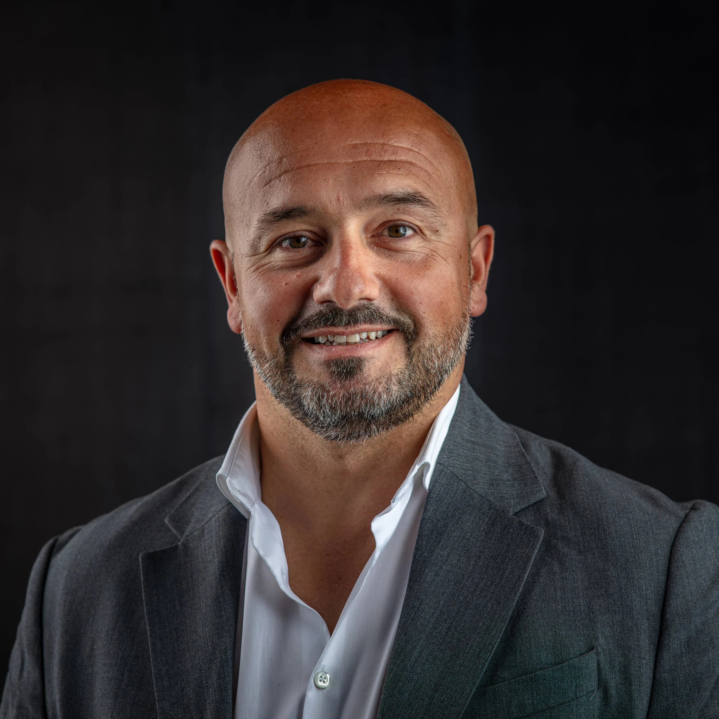 A middle-aged man with a beard, smiling, wearing a pink dress shirt and a dark blazer, posing indoors against a plain background.