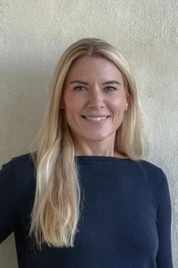 A smiling blonde woman with long hair, wearing a dark long-sleeve shirt, standing against a light-colored wall.