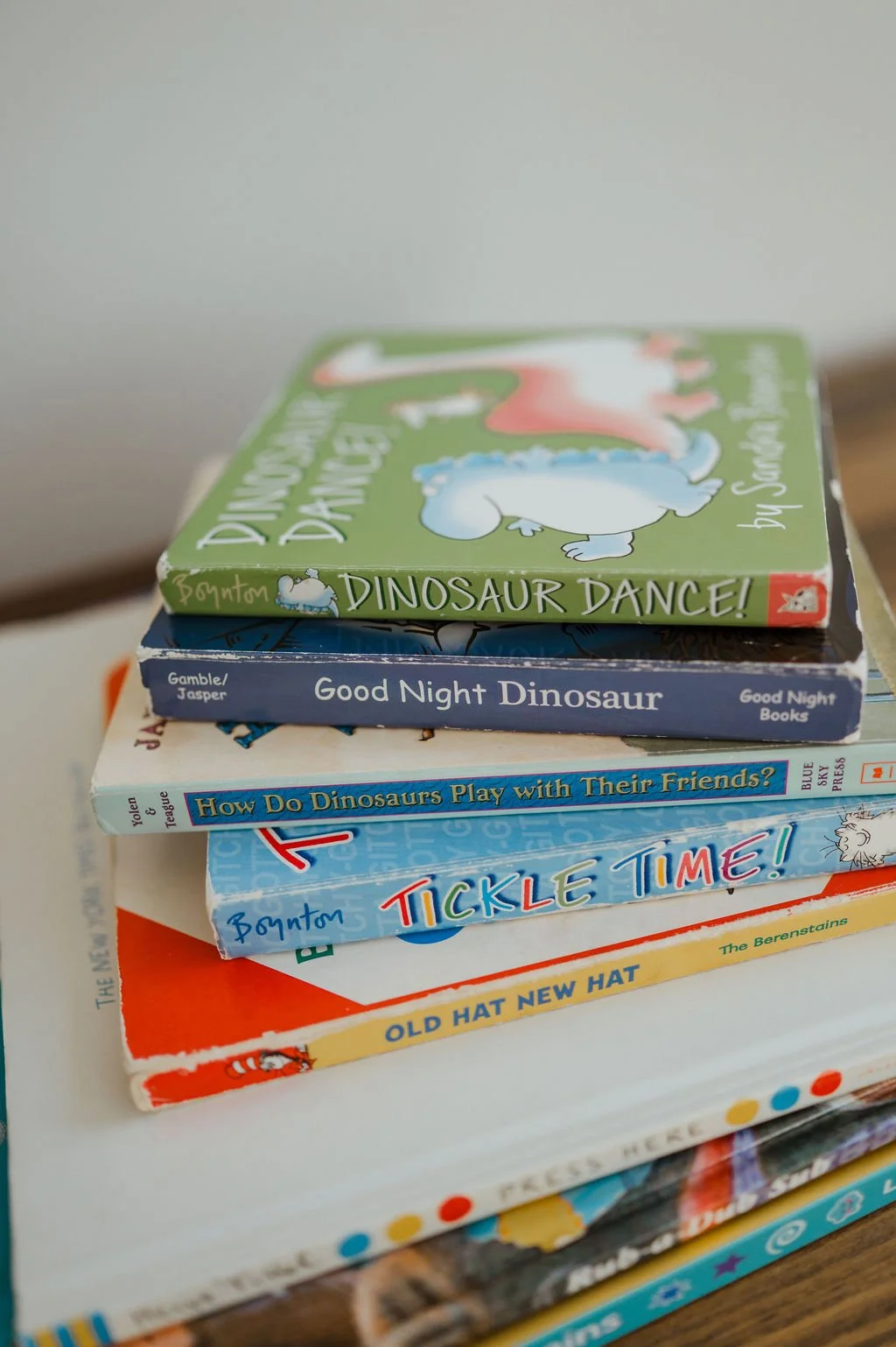 Stack of children's books featuring dinosaur themes, including titles like 'Dinosaur Dance!', 'Good Night Dinosaur', 'How Do Dinosaurs Play with Their Friends?', 'Tickle Time!', and 'Old Hat New Hat'.