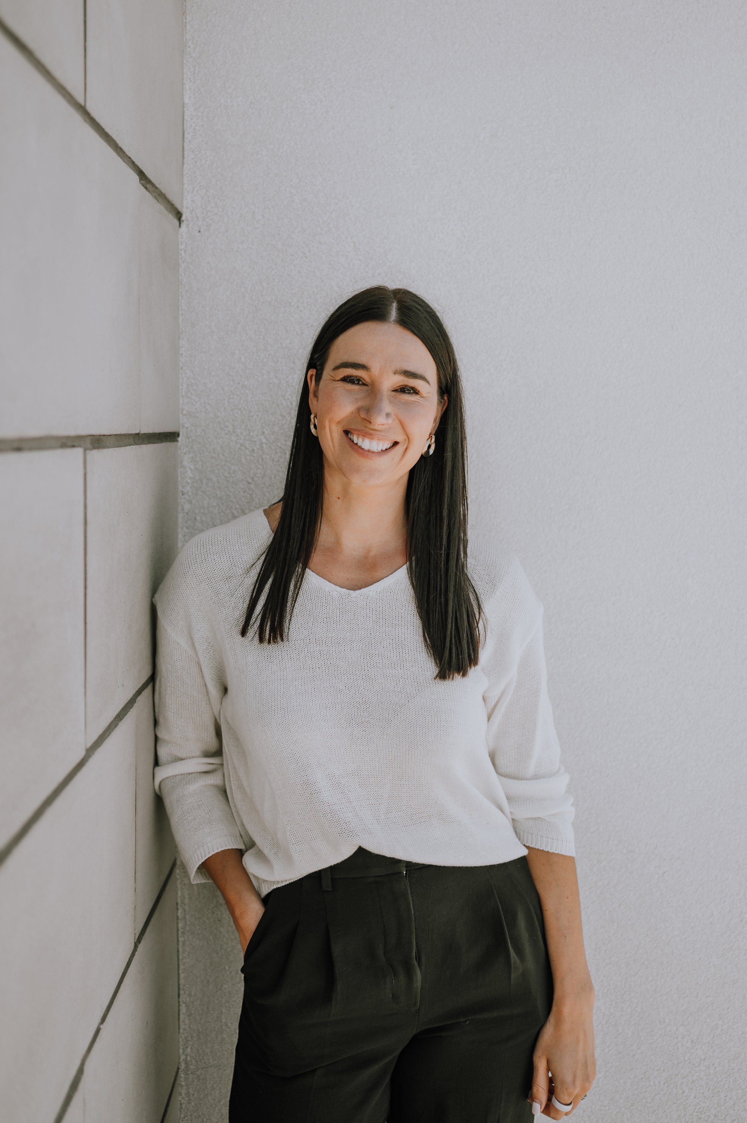 Young woman with long dark hair, smiling, wearing a black long-sleeve top and black pants, standing against a plain white background.
