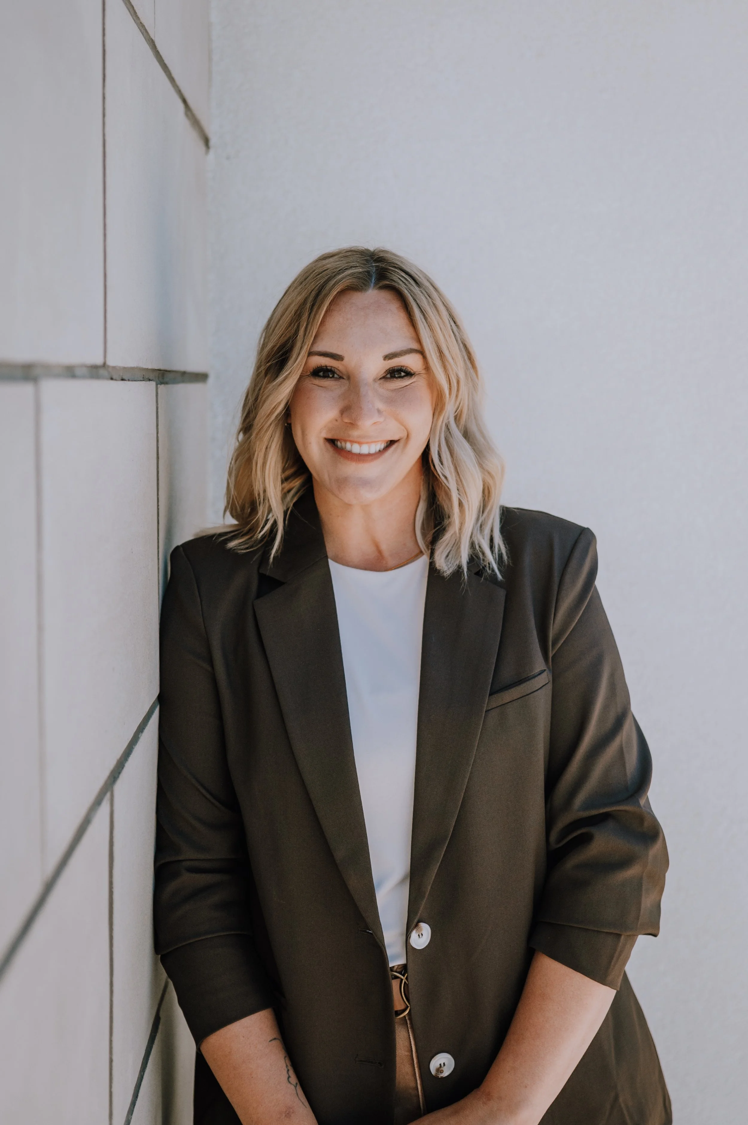 A smiling woman with shoulder-length wavy blonde hair, wearing a sleeveless white top and dark green pants, standing against a plain light background.